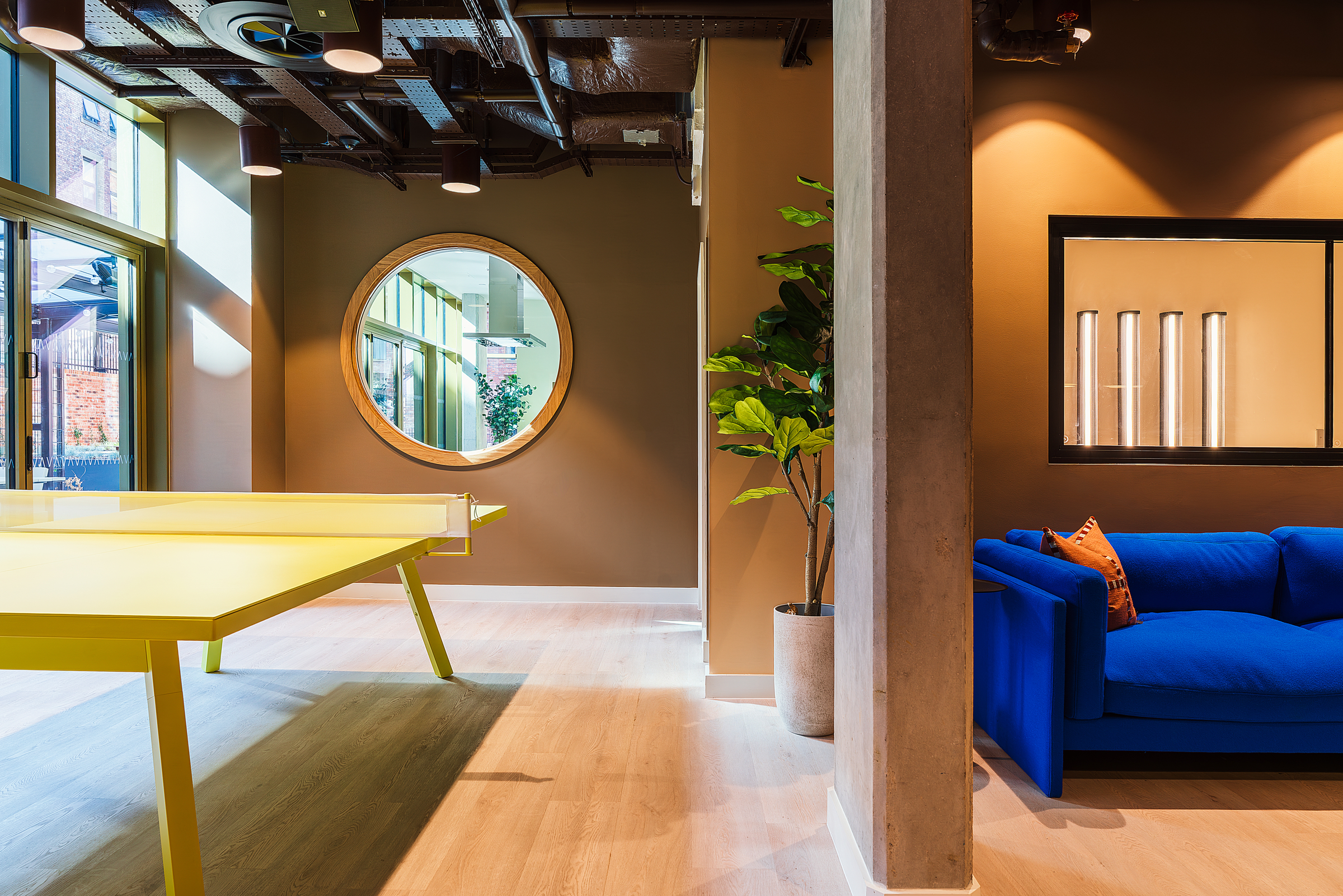 Modern interior with a yellow ping pong table, large round mirror, potted plant, and blue sofa. Warm lighting and a window create a cozy, inviting atmosphere.