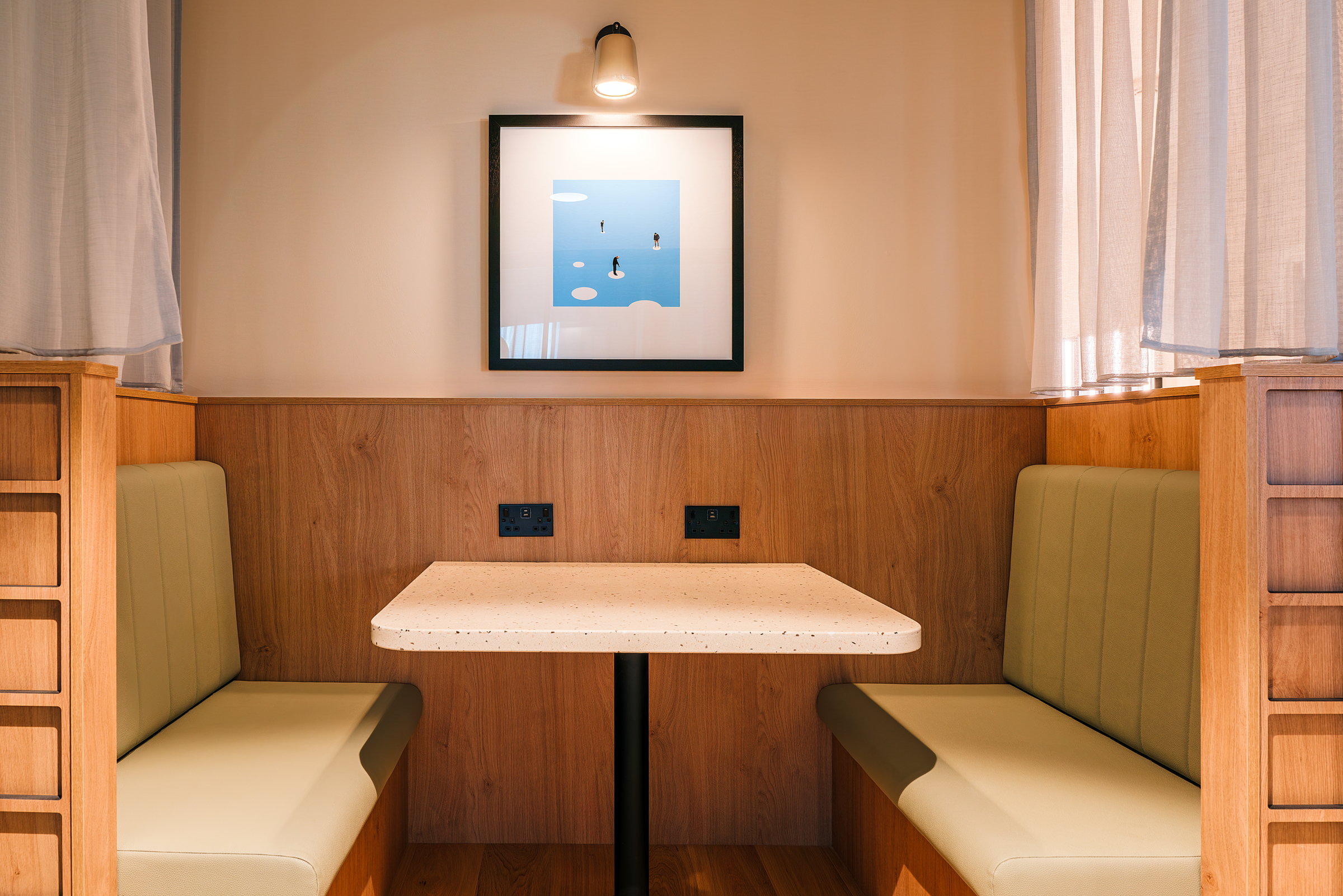 A cozy booth with light green cushioned seats, a terrazzo table, wood paneling, and a minimalist framed artwork on the wall under a small light, softly lit by daylight through curtains.