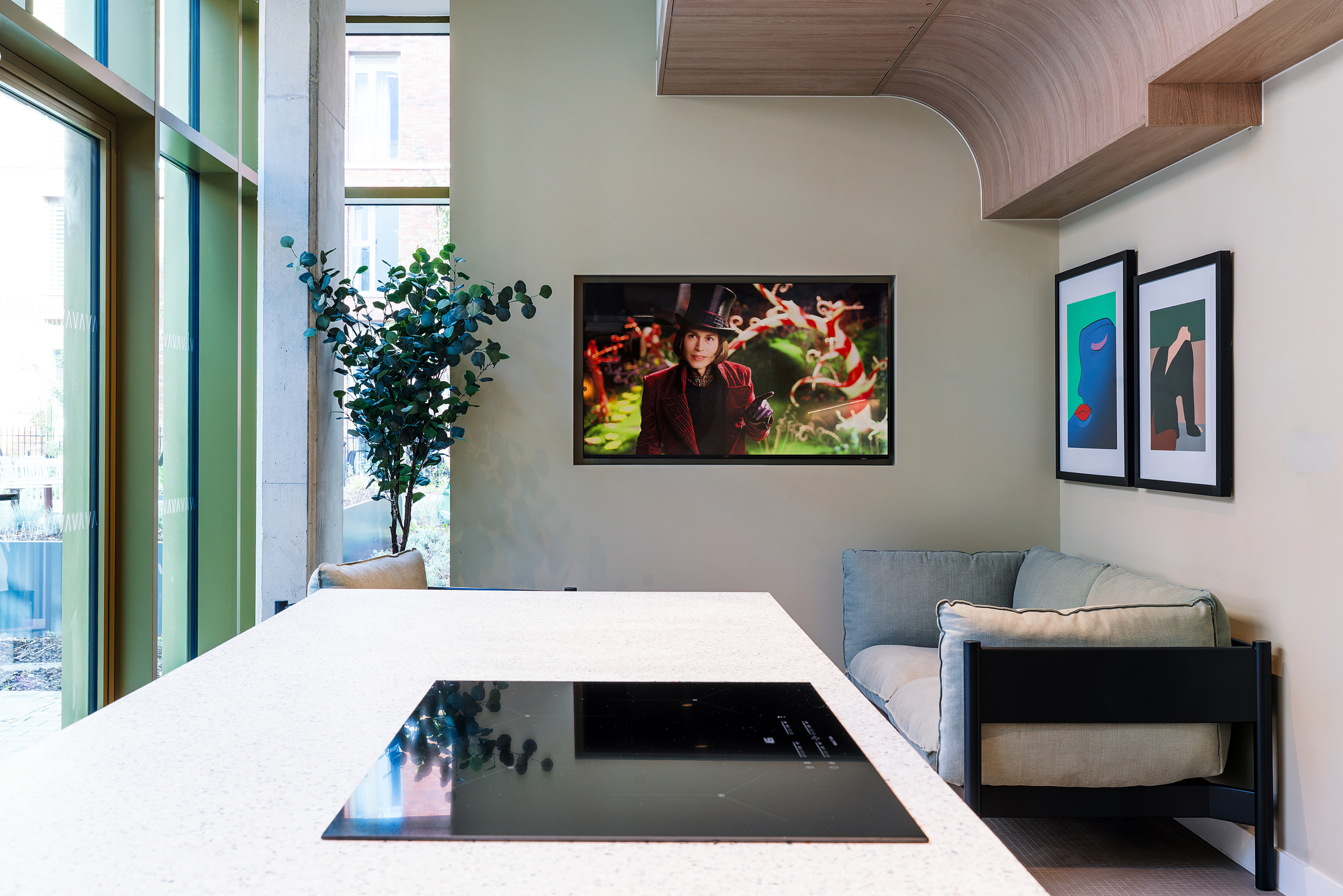 Modern lounge with cream sofa, framed art on the wall, and a TV displaying a scene from “Willy Wonka.” Large windows let in natural light, and a potted plant sits by the corner. Countertop in foreground.