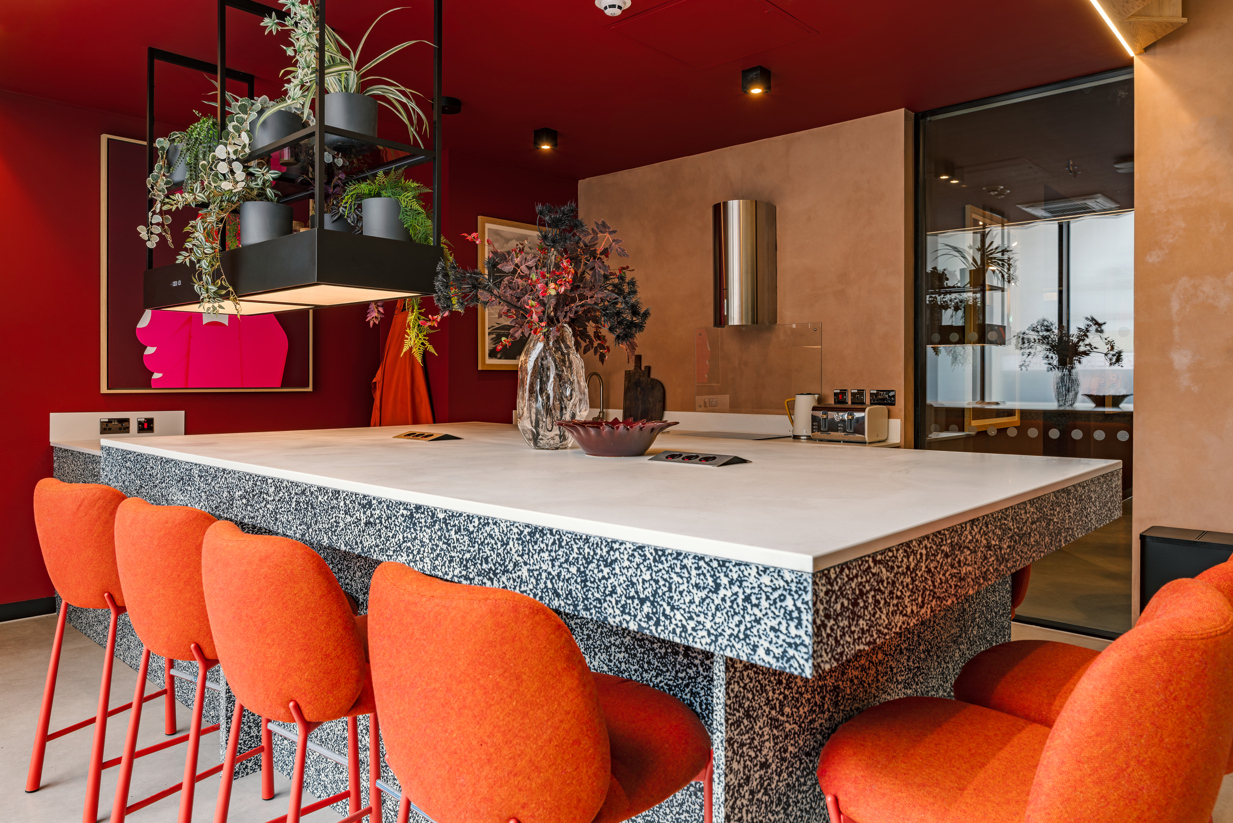 A modern kitchen at Vita Student features a terrazzo island, orange bar stools, hanging plants, and bold red walls. Contemporary decor includes artwork, vases, and sleek fixtures.