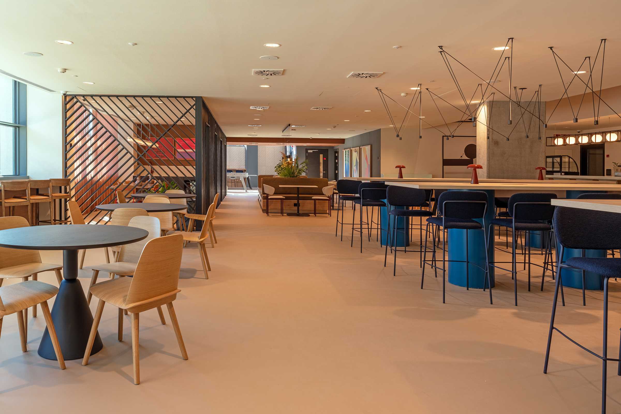 Modern open office lounge with tables, wooden chairs, high blue stools, a long counter, and decorative lighting. Wooden partition and artwork are in the background. Large windows let in natural light.