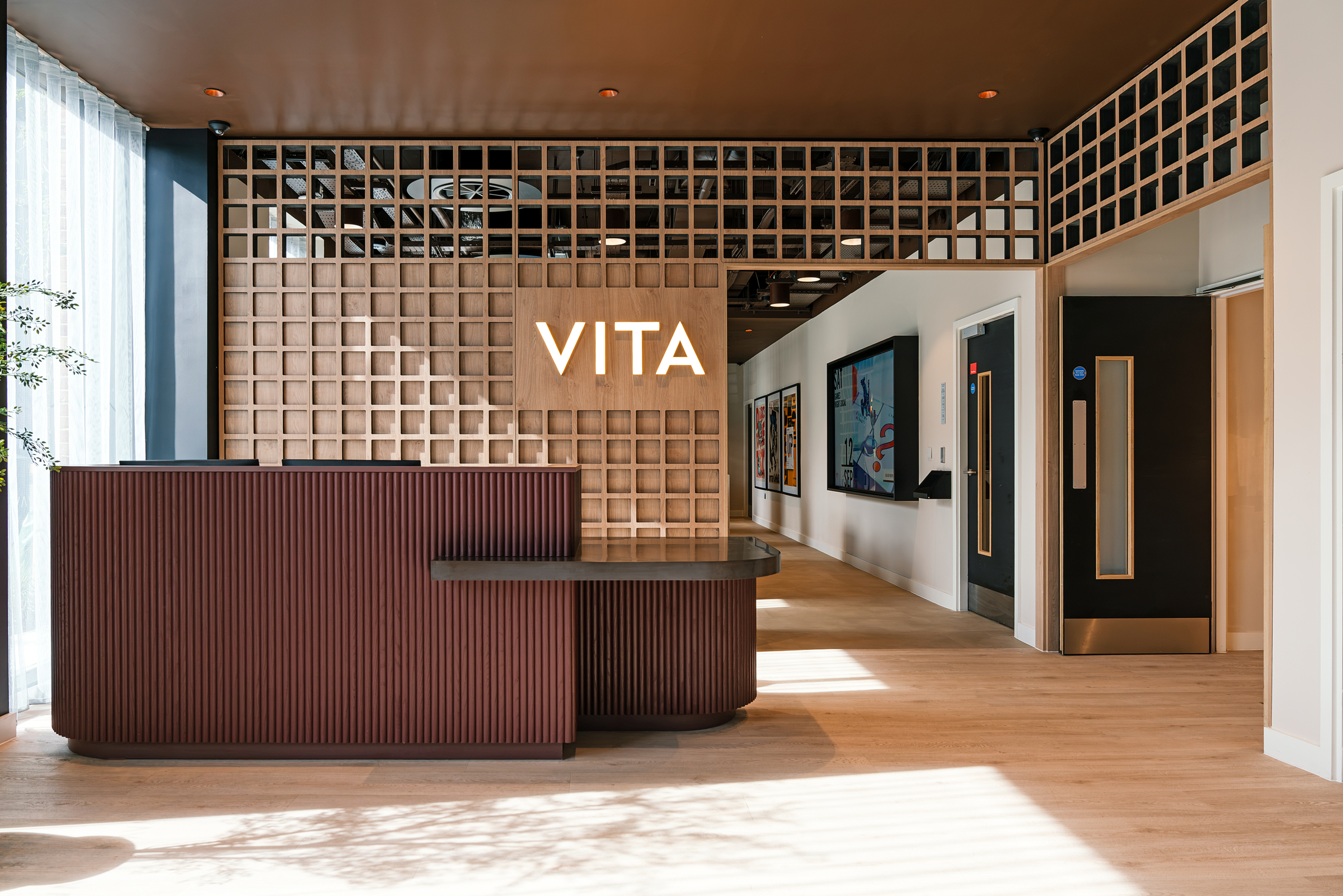 Modern reception area with a brown wood desk, geometric wall paneling, and the illuminated word “VITA” at the center. Perfect for welcoming guests on an open day, a bright hallway extends to the right, lined with artwork and doors.