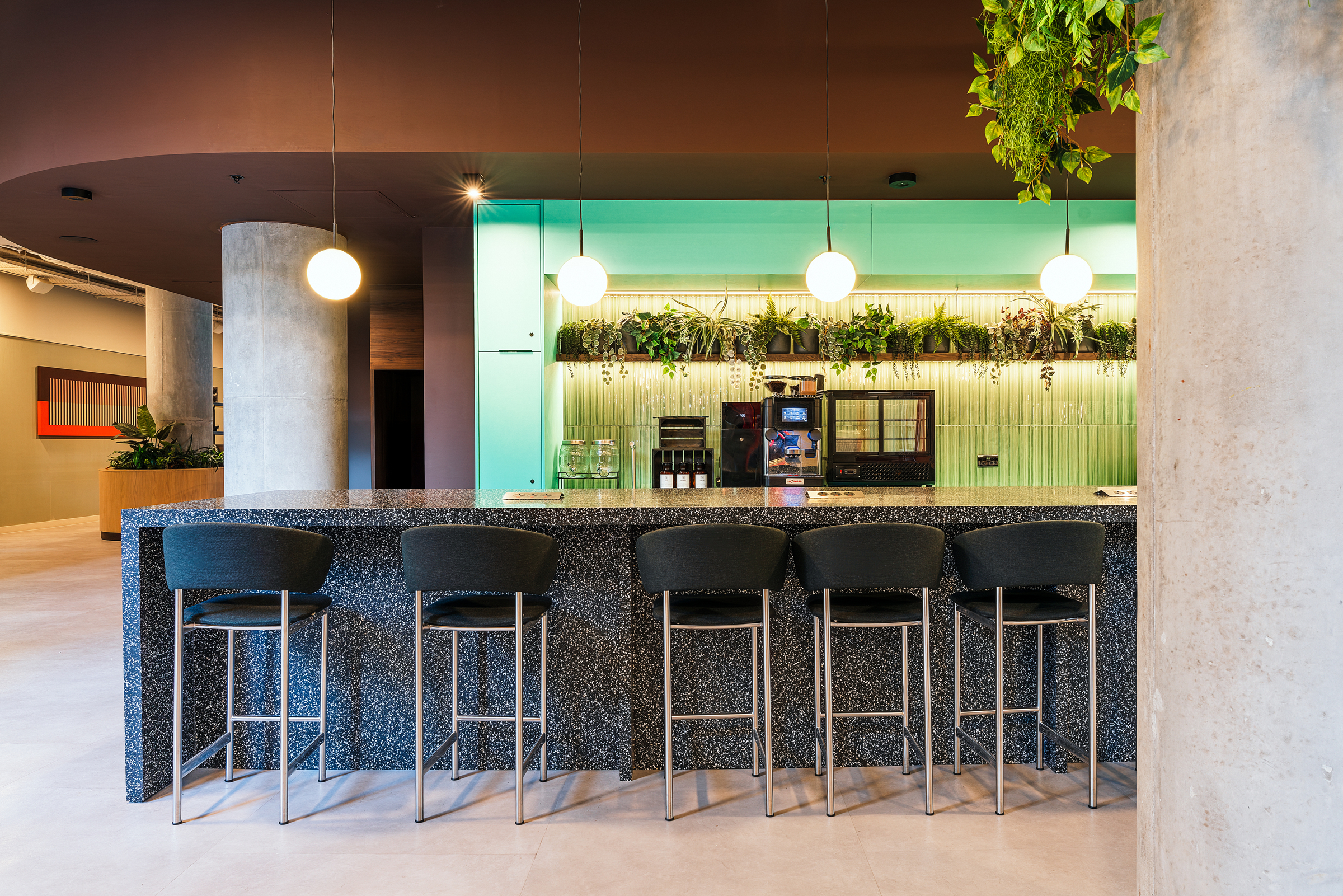 Modern Vita Student kitchen area with a long black island, five high chairs, hanging globes, a coffee machine, a microwave, and green plants above a backlit turquoise backsplash. A concrete pillar is visible on the right.