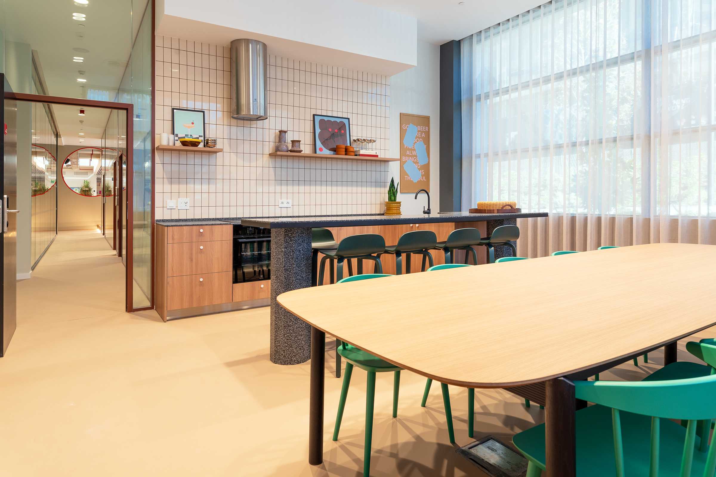 Modern kitchen with a long wooden dining table, green chairs, tile backsplash, open shelves, and large windows with sheer curtains letting in natural light.