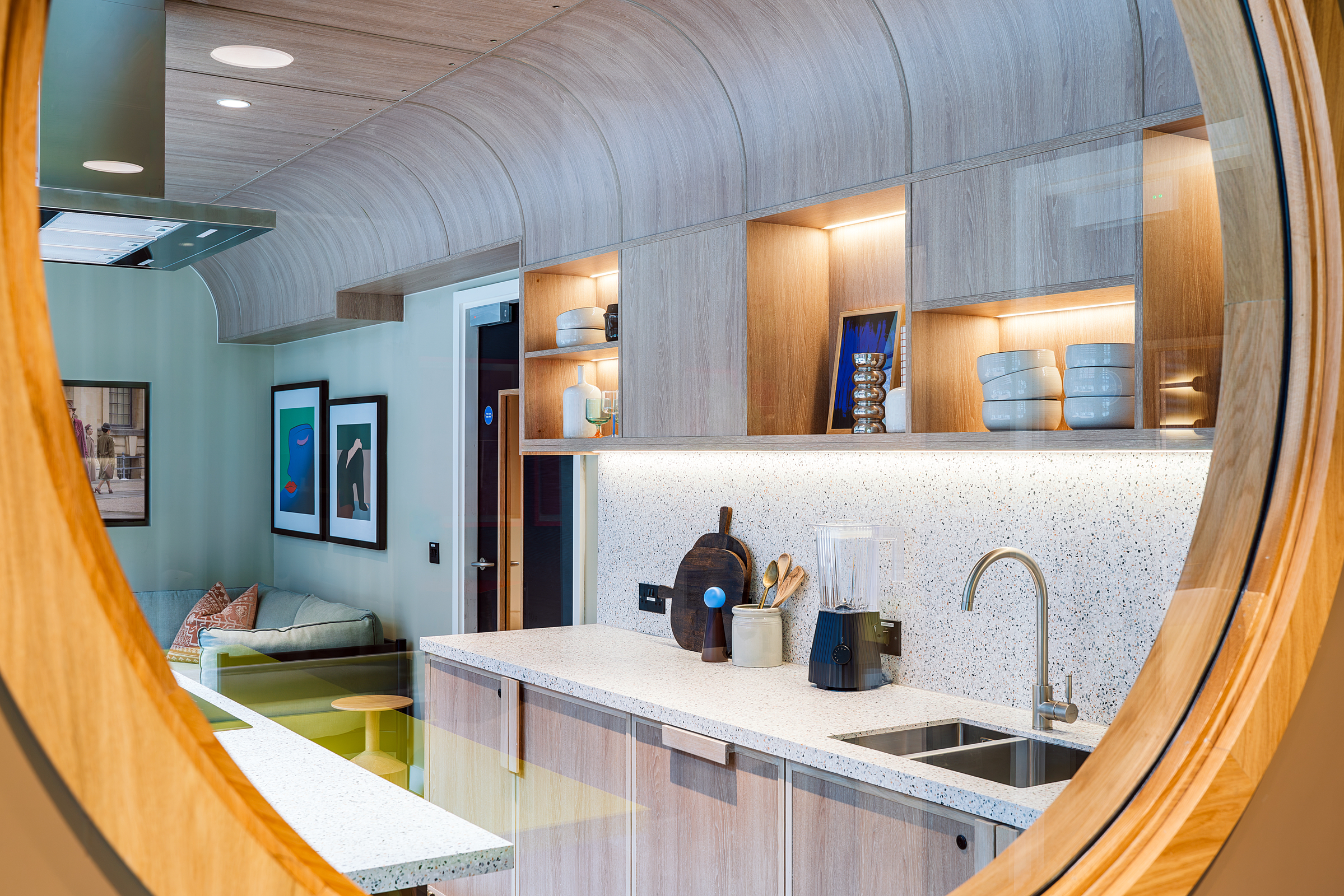 A modern kitchen with wood cabinets, open shelves, a sink, a blender, and decorative ceramics is beautifully displayed through a large round window from an adjacent room with green walls and artwork—creating an inviting atmosphere perfect for an open day.