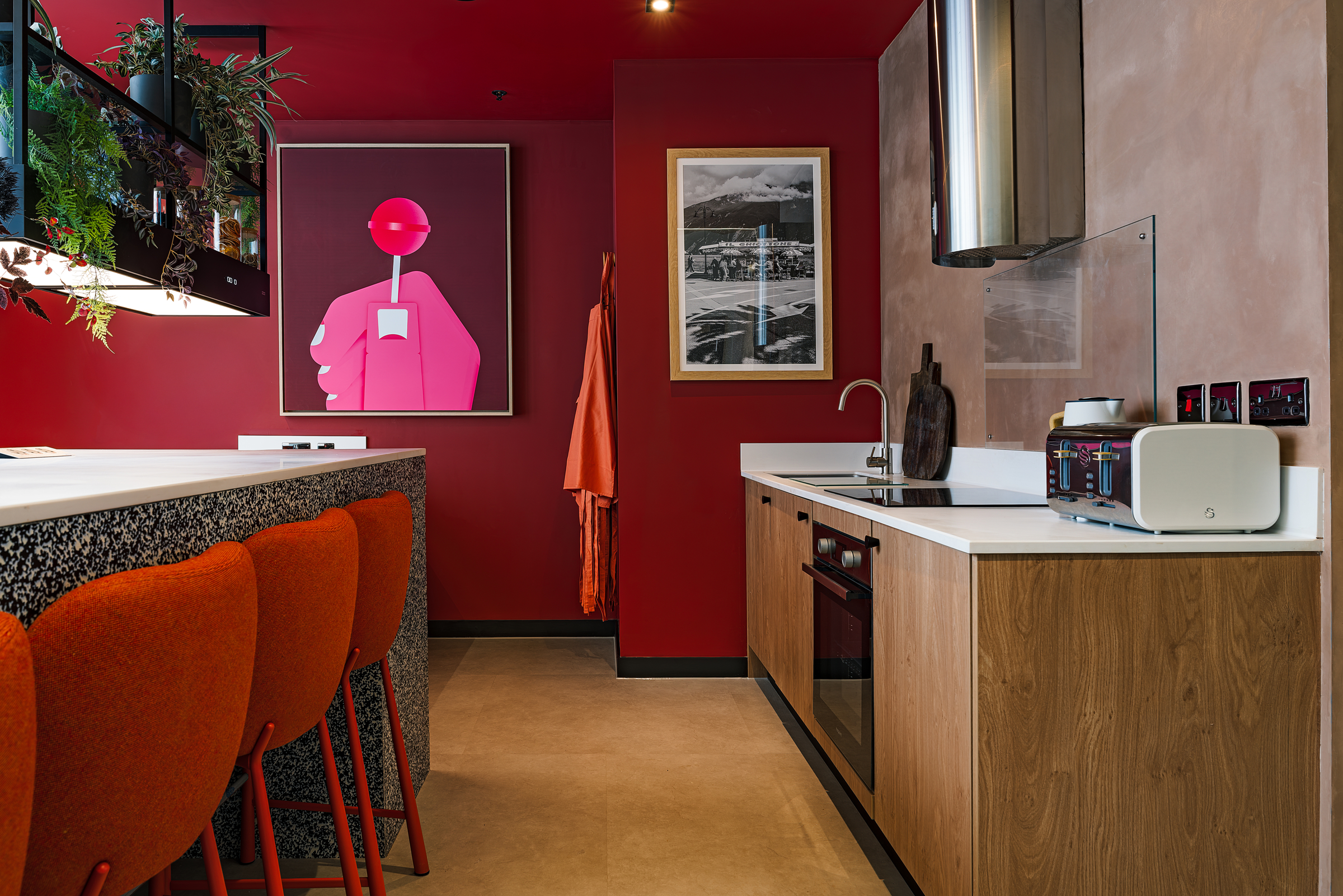 Modern kitchen with bold red walls, orange chairs at a breakfast bar, white countertops, wood cabinetry, potted plants, and colorful artwork on the wall—perfectly capturing the vibrant atmosphere of Vita Student living. Appliances and utensils are neatly arranged on the counters.
