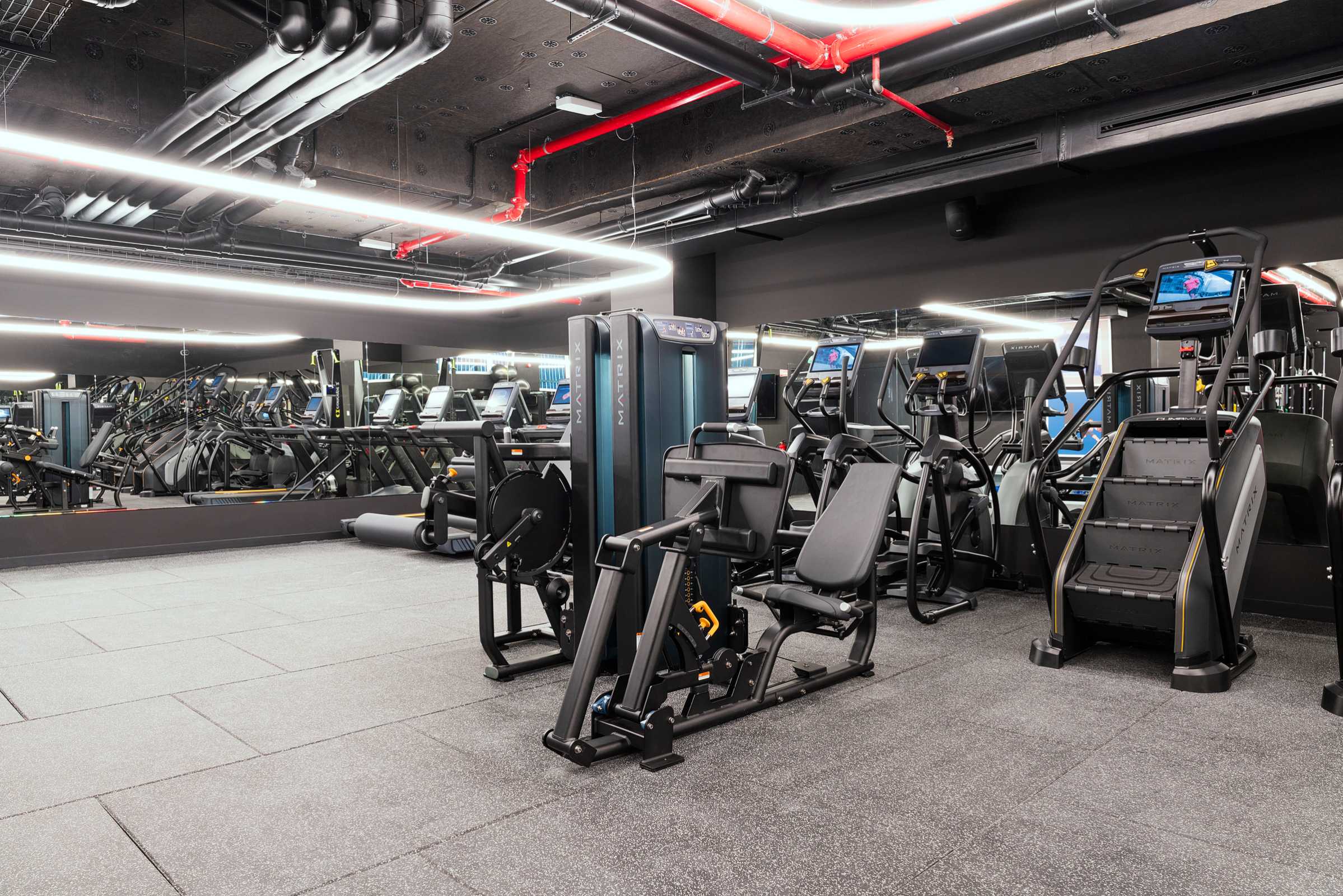 A modern gym with exercise machines, including a stair climber, a seated leg press, and treadmills, all set on gray flooring with large mirrors and overhead lighting.