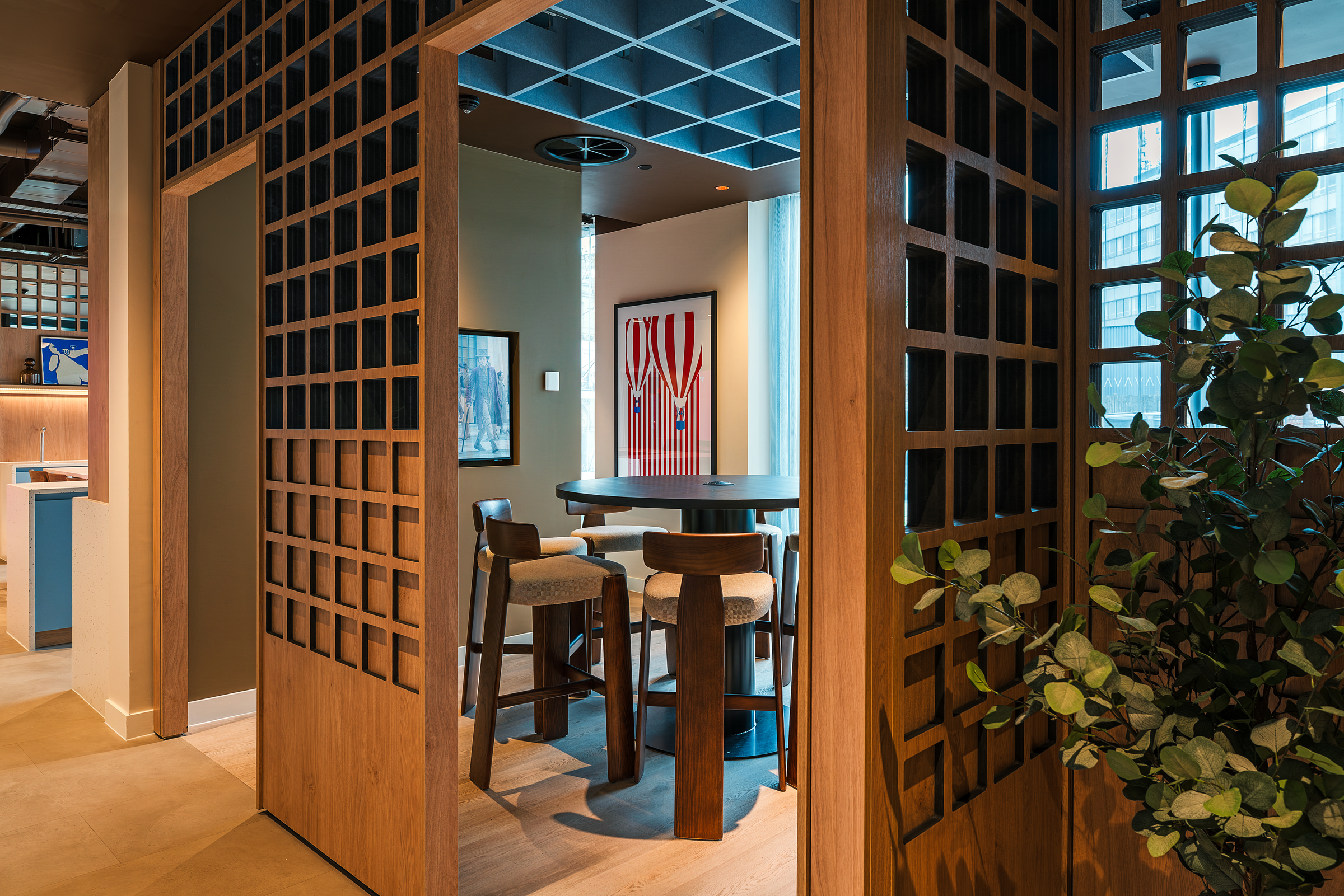 A modern office lounge designed for open day visitors, featuring wooden grid partitions, a round table, four cushioned chairs, a potted plant, wall art, and large windows allowing natural light to brighten the space.
