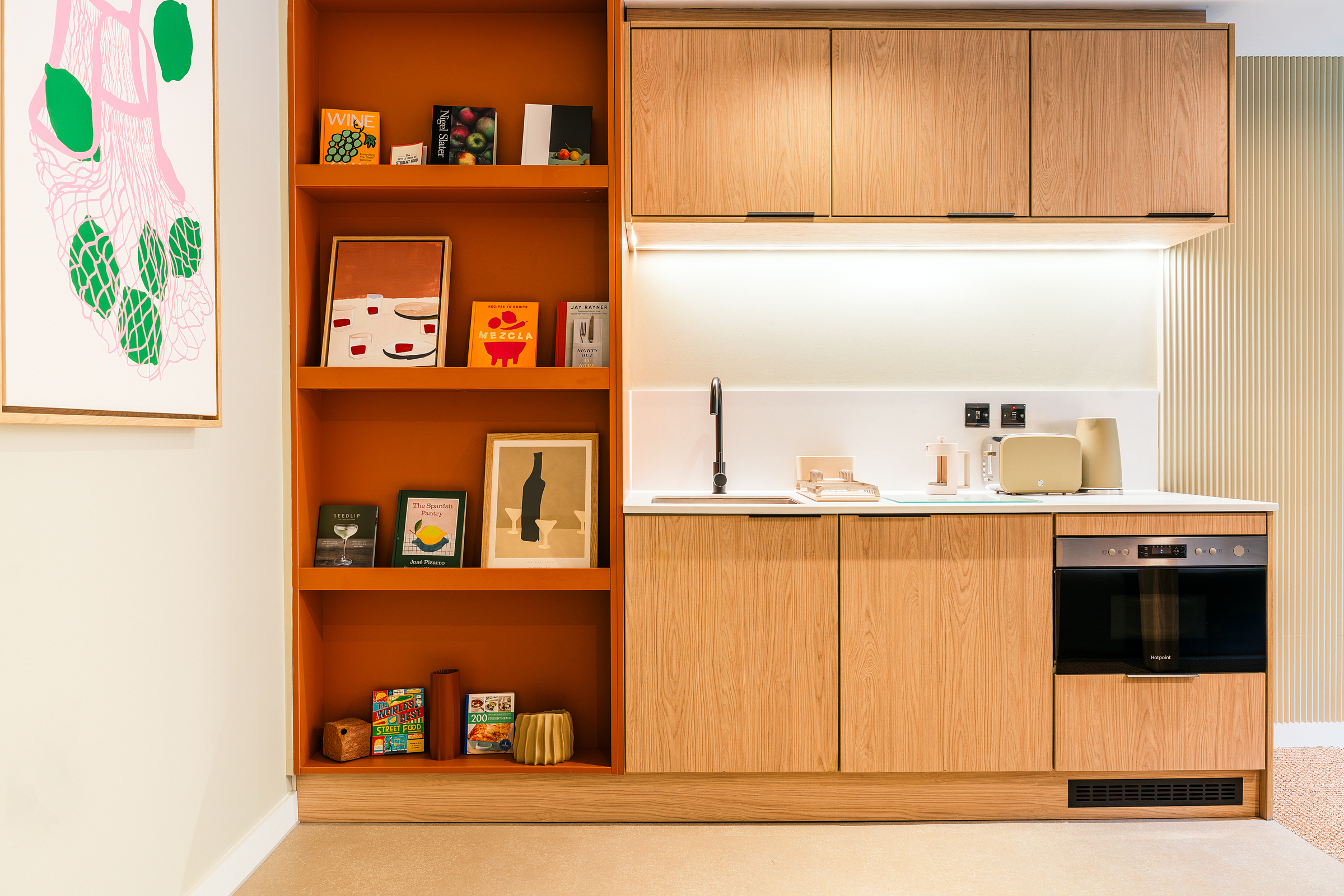 A modern Vita Student kitchenette with light wood cabinets, open orange shelves displaying art and books, a built-in oven, sink, toaster, and a framed abstract artwork hanging on the adjacent wall.