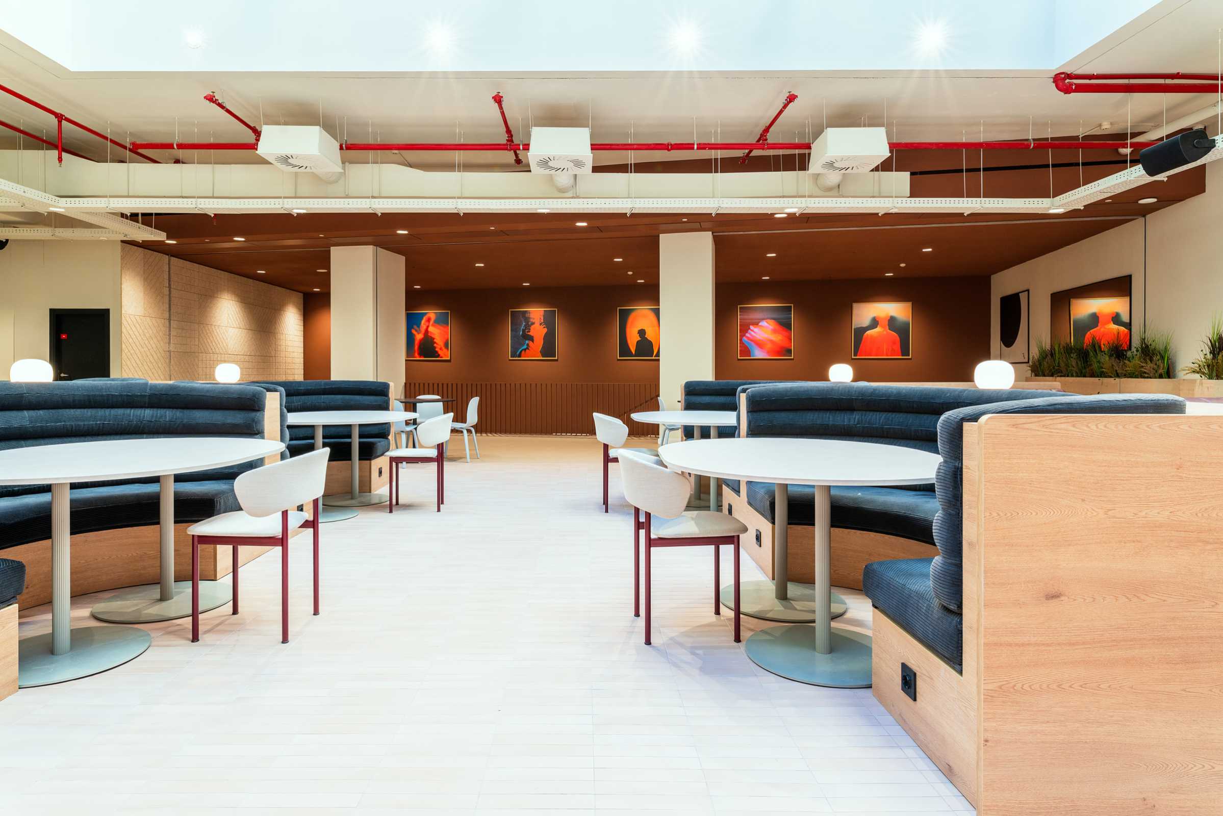 A modern, well-lit dining area with round tables, curved booth seating, white chairs, and abstract artwork on the brown wall in the background. Red ceiling pipes and globe lights are visible throughout the space.