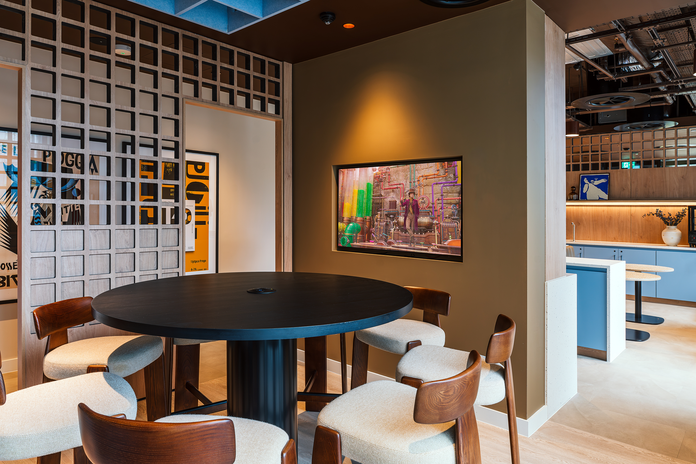 Modern office lounge with a round black table, five cushioned wooden chairs, wall art, decorative lattice, and a kitchenette in the background with blue cabinets and a countertop—an inviting space perfect for an open day event.