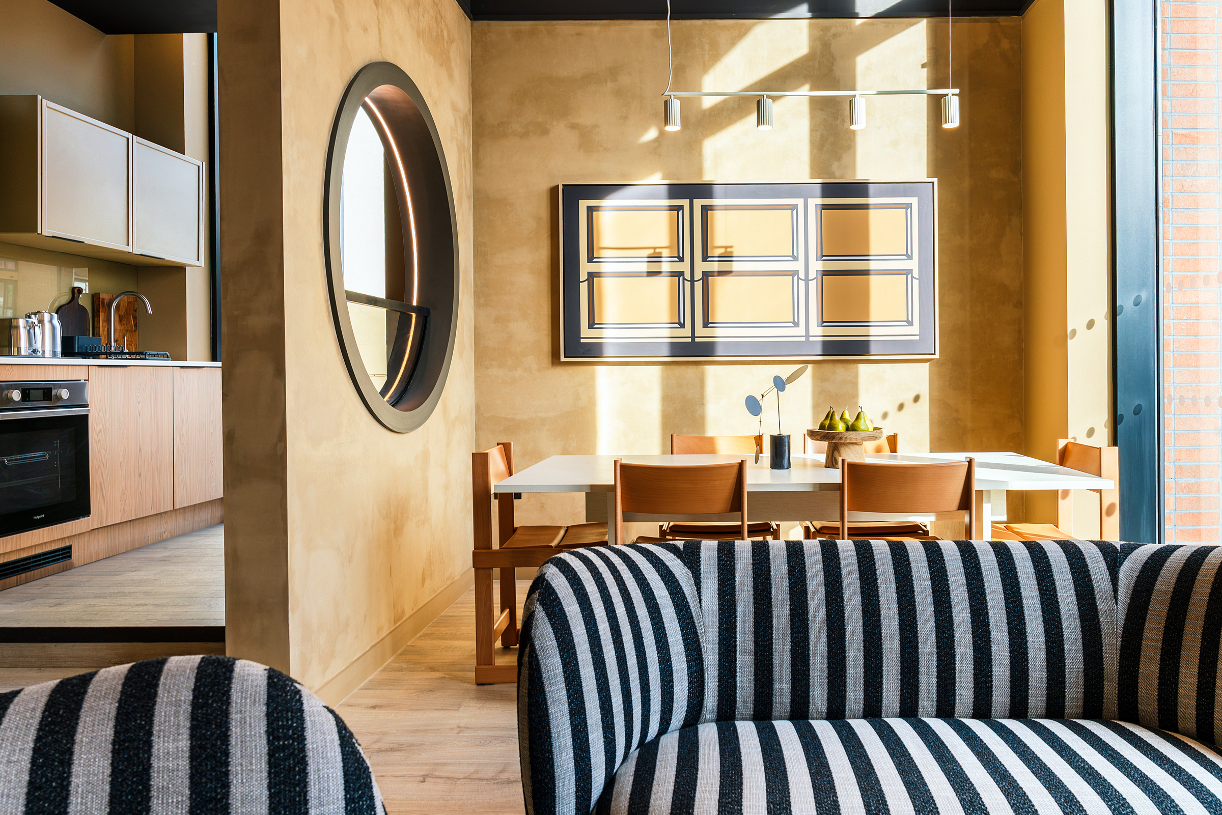 Modern dining area with a rectangular table, wooden chairs, striped sofa in the foreground, round window, and abstract wall art—much like the inviting settings found at Vita Student accommodations. Warm sunlight streams in, highlighting beige walls and minimalist decor for a contemporary yet cozy atmosphere.