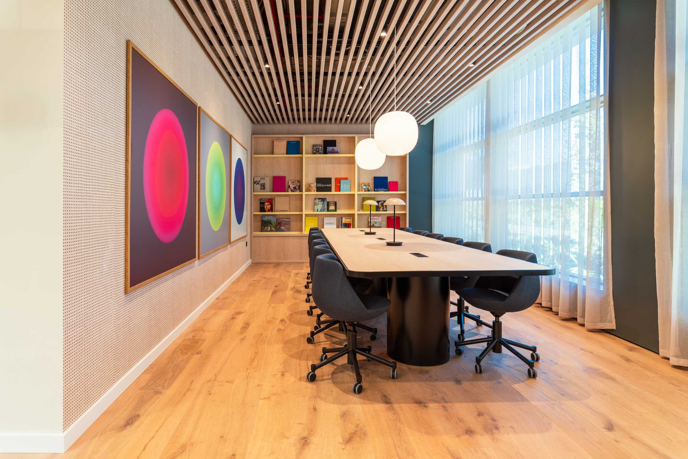 Modern conference room with a long table, black chairs, large colorful circular artwork on the wall, bookshelf with books, wooden floor, and large windows with sheer curtains letting in natural light.
