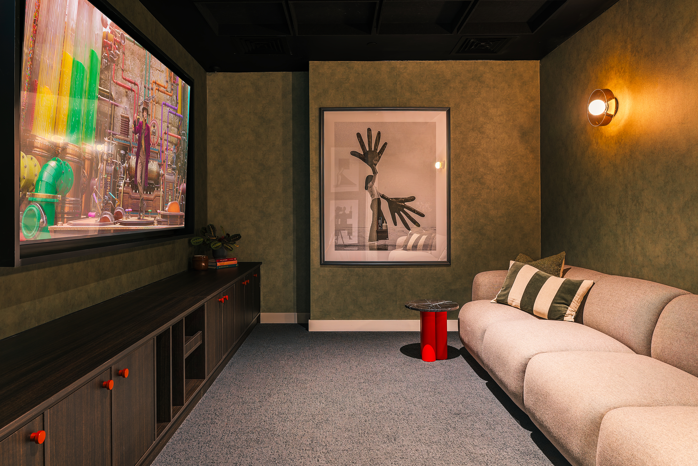 A cozy room welcomes you for the open day, featuring a large wall-mounted TV showing an animated film, a beige sofa with pillows, a small red side table, a modern light, and framed art of hands displayed on the green wall.