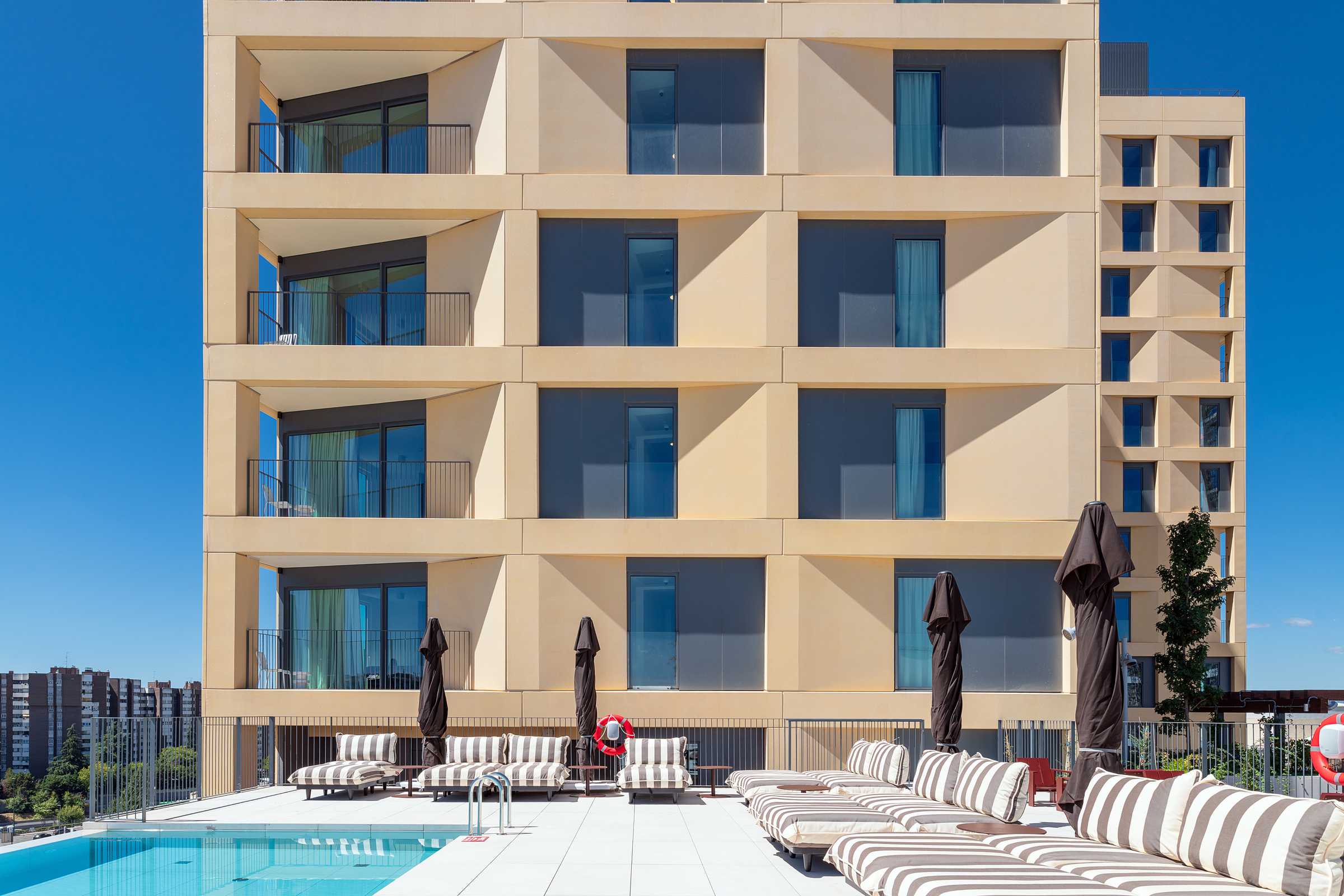 Modern apartment building with balconies behind a poolside area featuring striped lounge chairs and closed umbrellas under a clear blue sky.