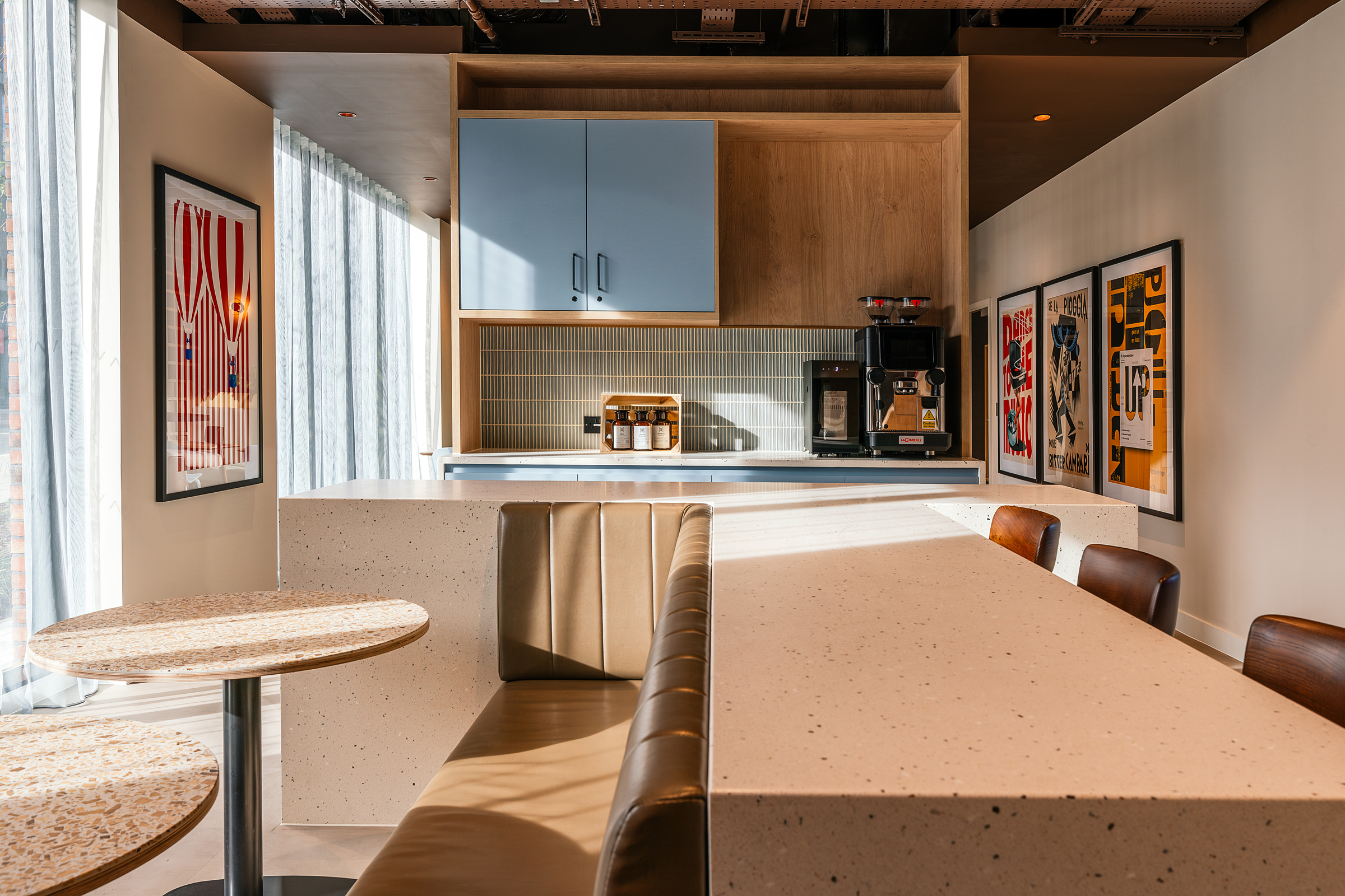 Modern cafe with a beige booth, round and rectangular tables, wall art, a coffee machine, and shelves. Sunlight streams through large windows with sheer curtains, creating a bright and inviting atmosphere—perfect for an open day visit.
