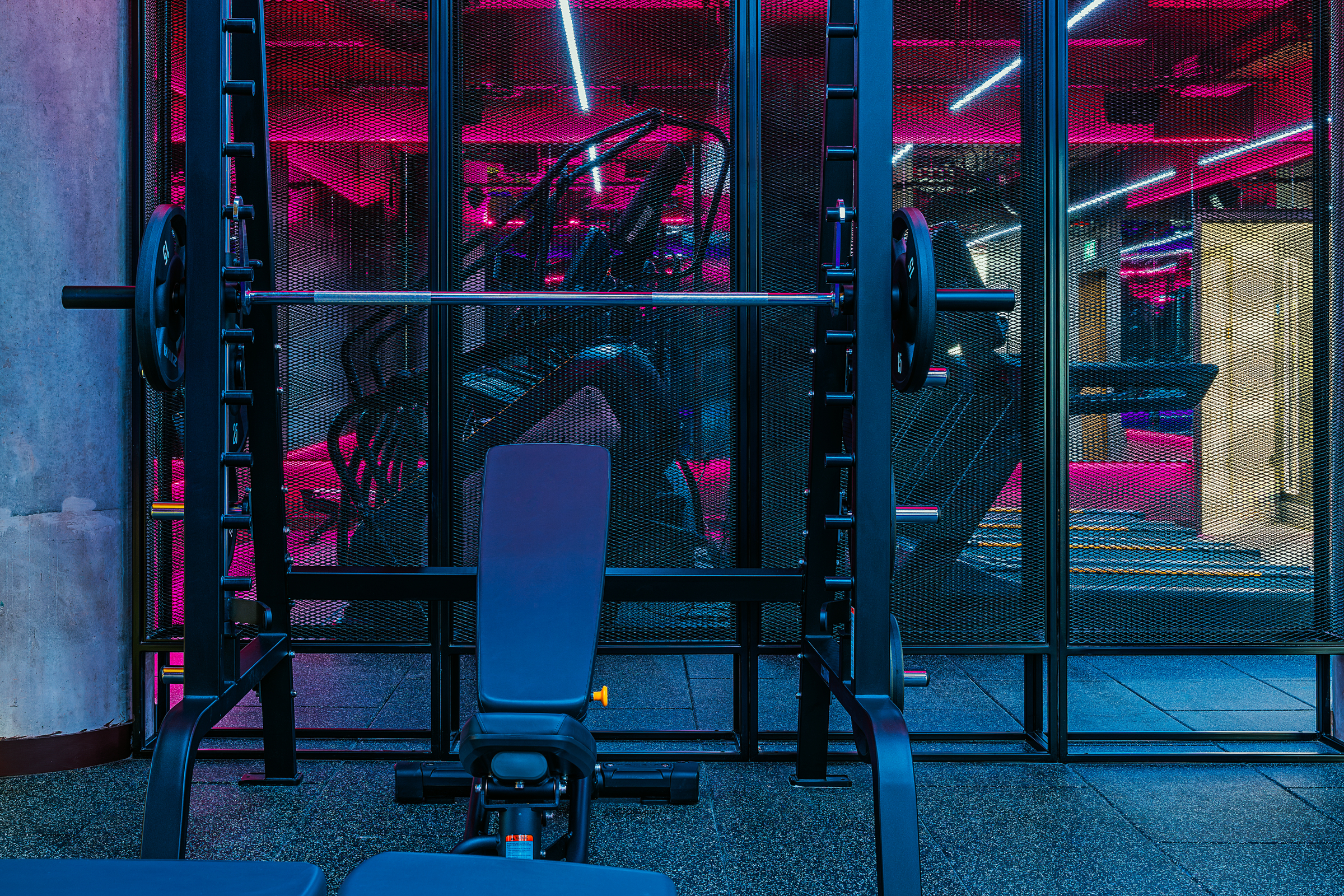 A bench press setup in a modern Vita Student gym with neon pink and blue lights, metal fencing, and exercise machines in the background.