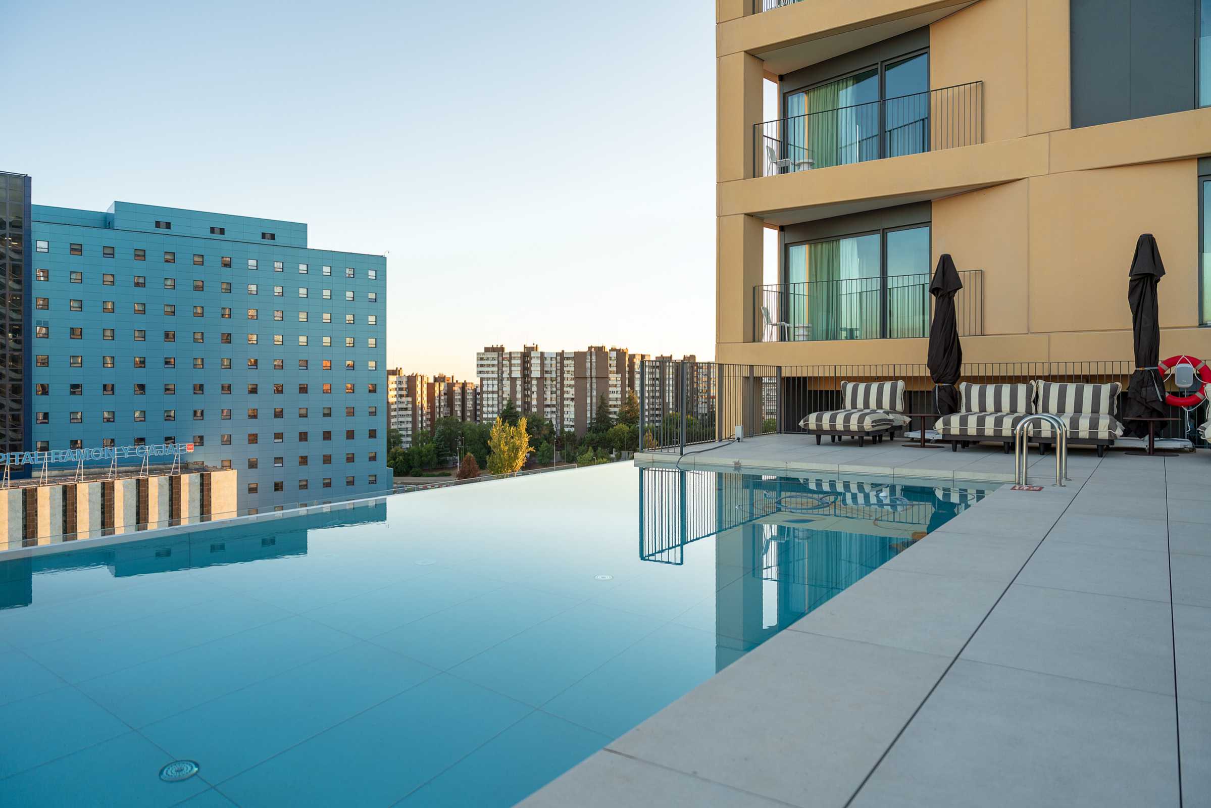 Rooftop infinity pool beside a modern building, with striped lounge chairs and closed umbrellas, overlooking a cityscape with tall buildings at sunset.