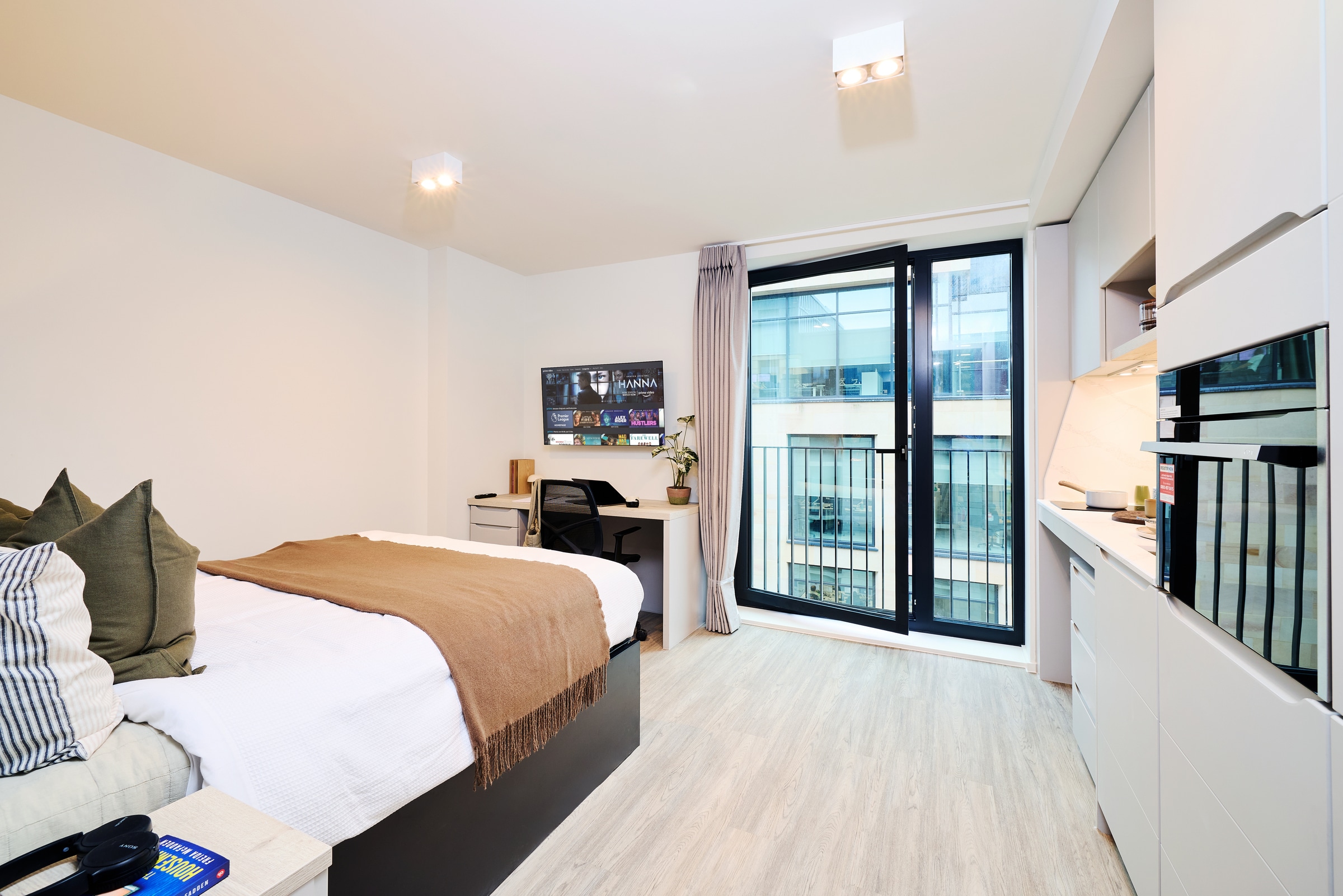 Modern studio apartment with a bed, desk, chair, TV, kitchenette, and large window with a balcony view. Light wood flooring and minimalist decor create a bright, cozy atmosphere.