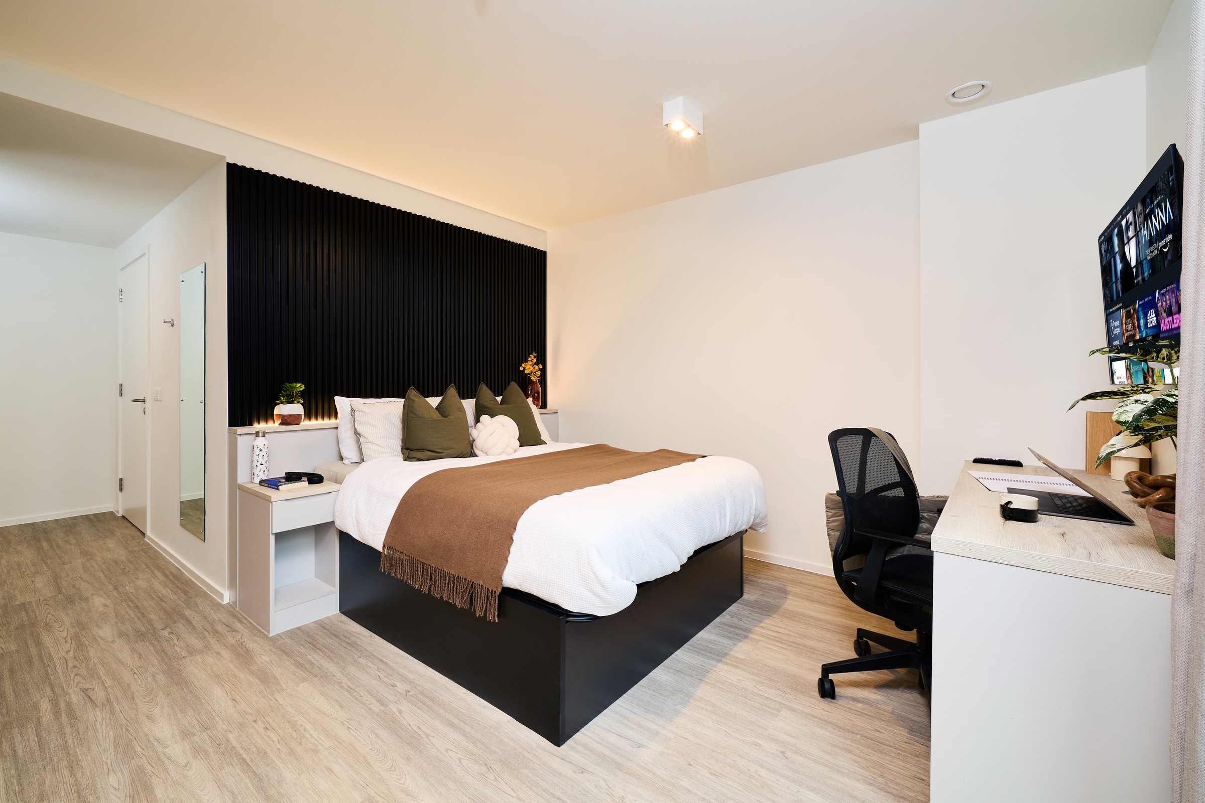 A modern bedroom with a double bed, brown blanket, and green pillows. A desk with a computer and chair sits to the right. Light wood flooring and white walls create a clean, minimalist look.