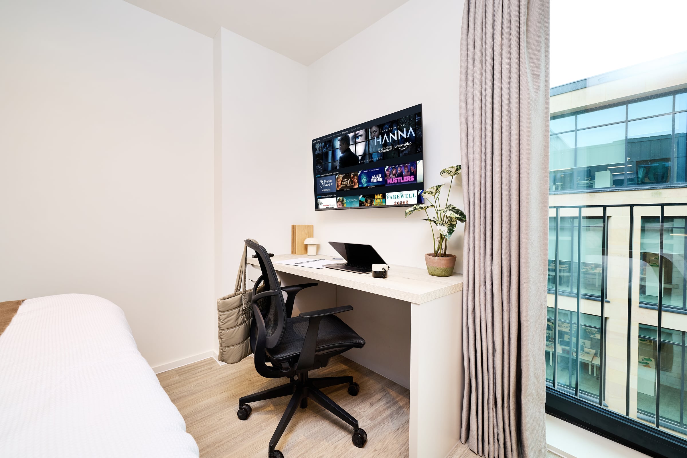Minimalist home office with a wall-mounted desk, black chair, laptop, potted plant, and TV displaying streaming apps. Floor-to-ceiling window shows a modern building outside. Light wood floor and white walls.