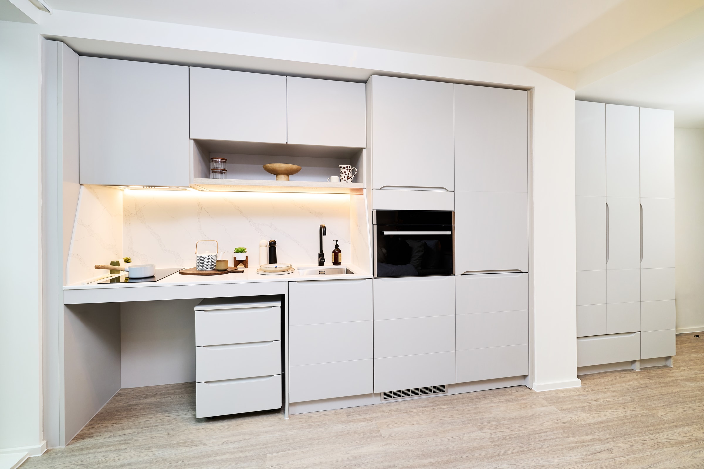 Modern kitchen with light gray cabinets, built-in oven, white marble backsplash, and light wood flooring. Open shelves display decor items; under-cabinet lighting brightens the countertop workspace.