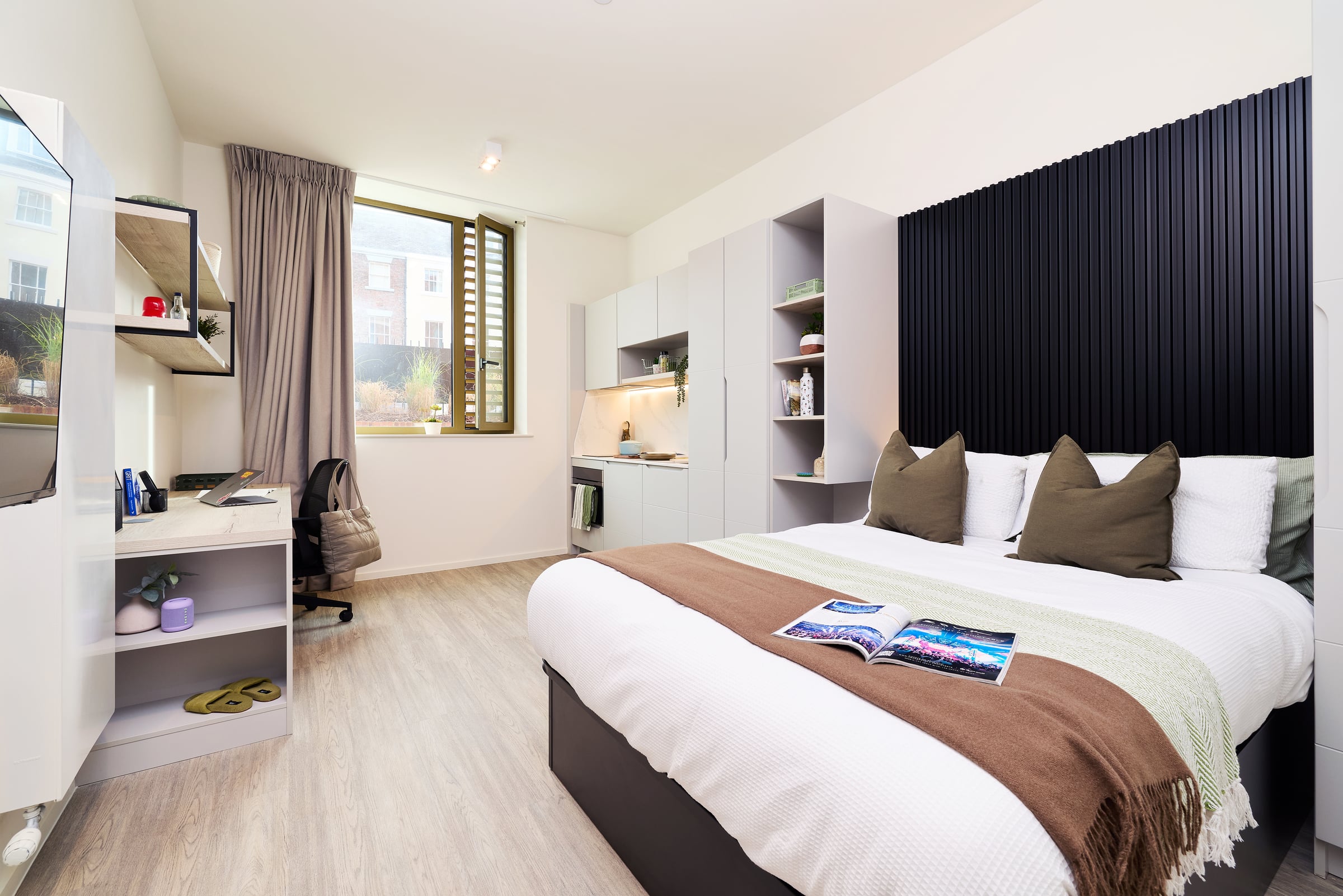 A modern bedroom with a large bed, brown blanket, and pillows. Desk with chair, laptop, and shelves; kitchenette and window with brown curtains. An open magazine rests on the bed. Light wood floor.