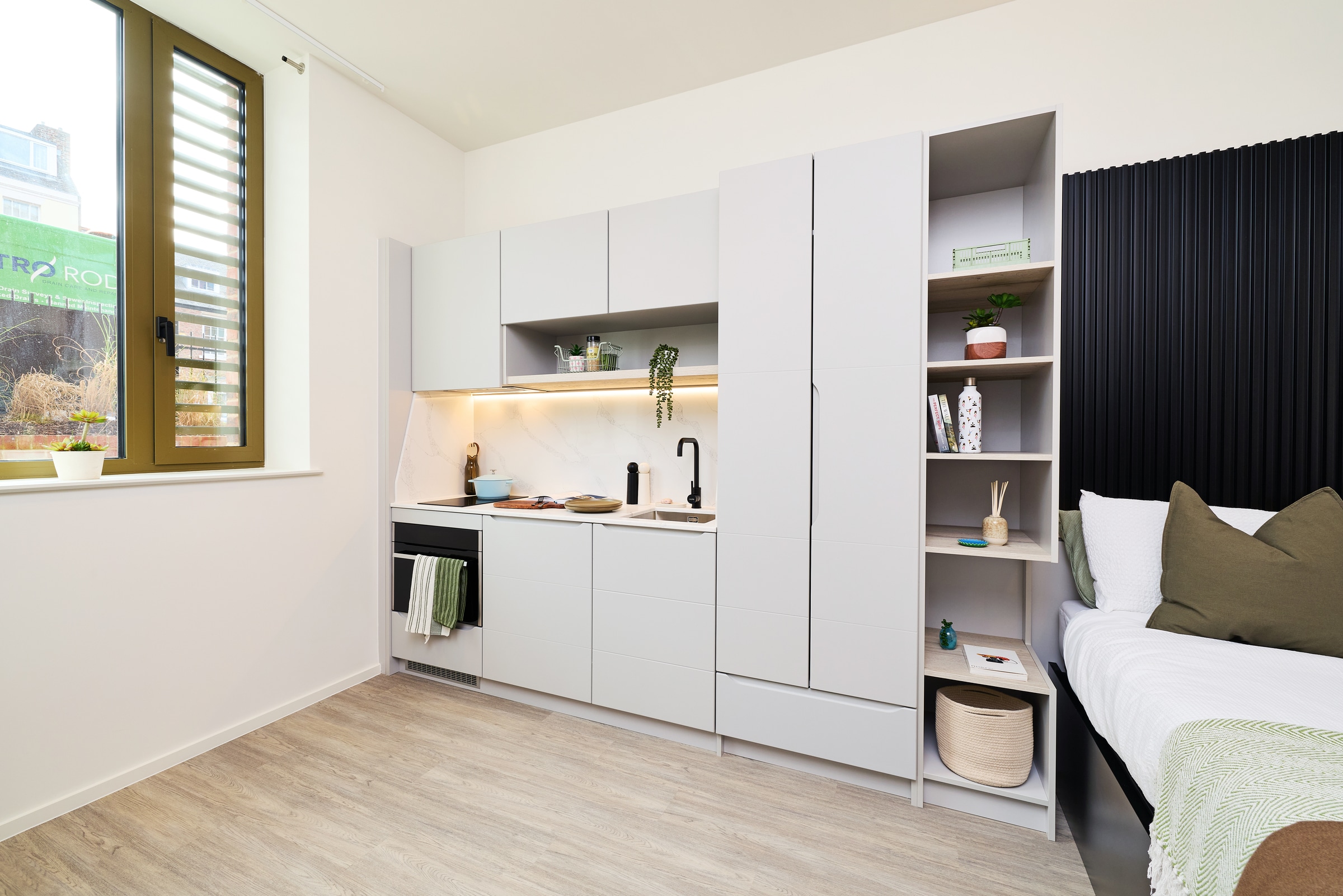 Modern studio apartment kitchenette with gray cabinets, sink, stove, and open shelving, next to a bed. A window lets in natural light. Minimalist decor includes plants, books, and simple accessories.