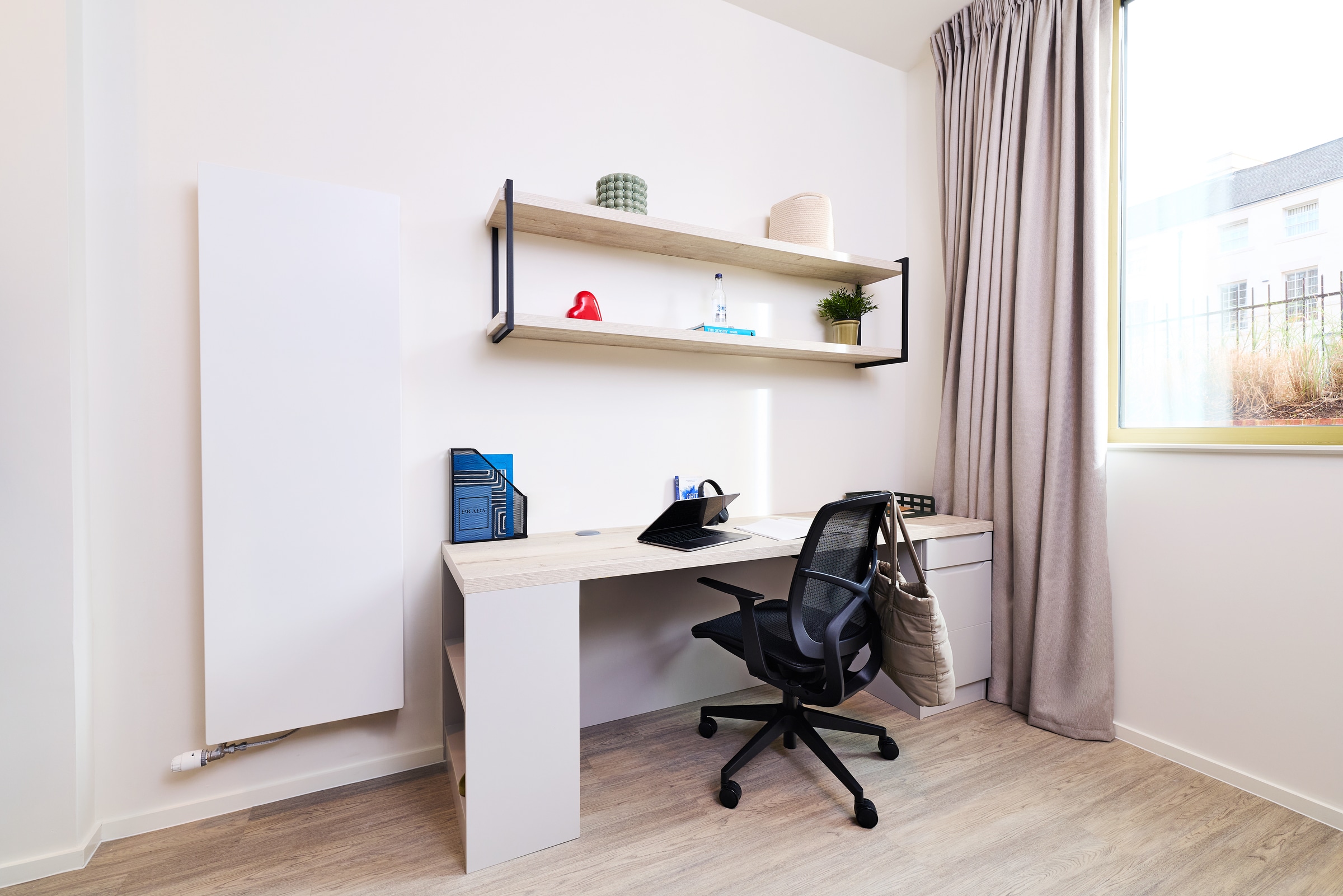 Minimalist home office with a desk, black chair, closed laptop, notebook, and desk organizer. Two shelves hold decor items, a water bottle, and a small plant. Large window with beige curtains lets in natural light.