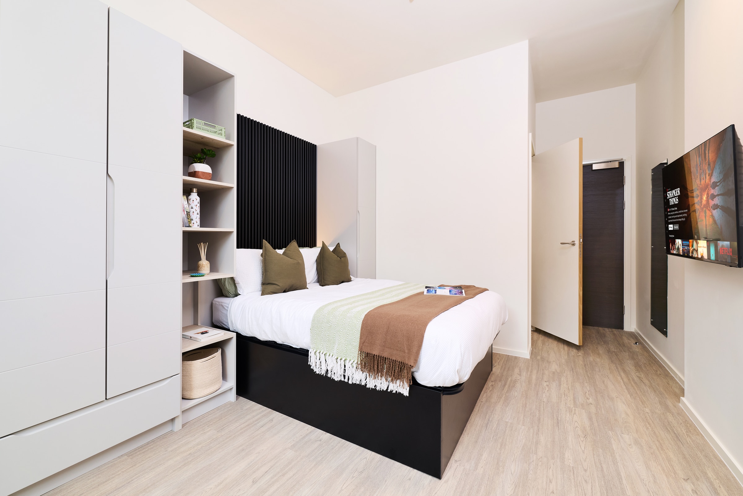 Modern bedroom with a neatly made bed, green and brown throw, open shelving with decor items, mounted TV on the wall, and wooden floor. The room is bright with white walls and minimalistic furniture.