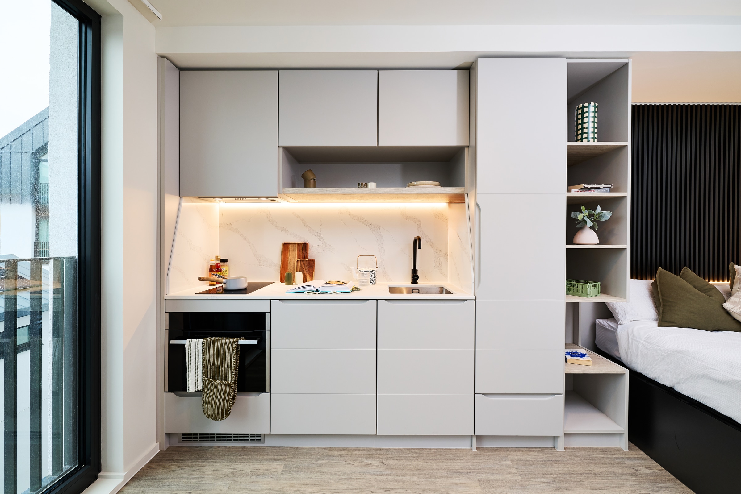 Modern studio kitchenette with light gray cabinets, built-in oven, cooktop, sink, open shelving, and a bed nearby. Warm under-cabinet lighting highlights countertop items and marble backsplash. A large window is on the left.