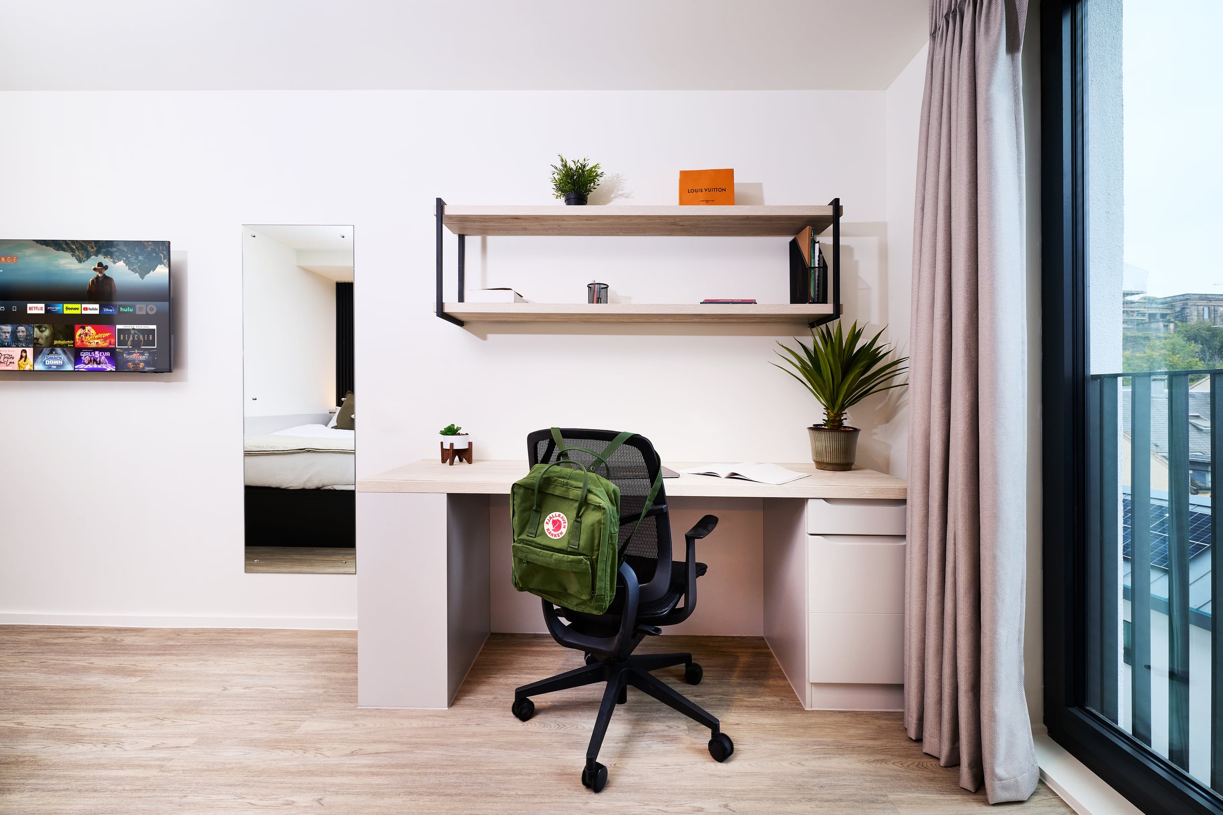A modern desk with a chair holding a green backpack, shelves with books and plants, near a window with curtains. A bed and TV are visible in the background.