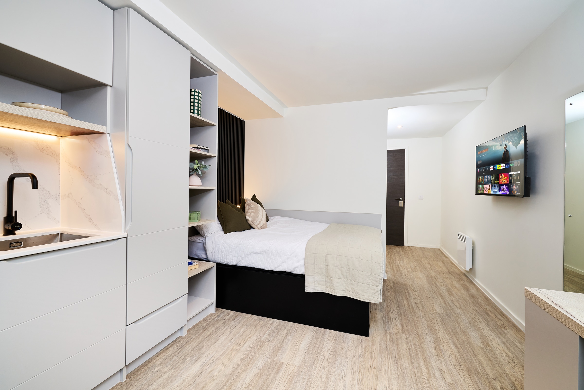 Modern studio apartment with a bed, open shelving, small kitchen area on the left, light wood floors, a beige blanket, wall-mounted TV, and neutral decor.