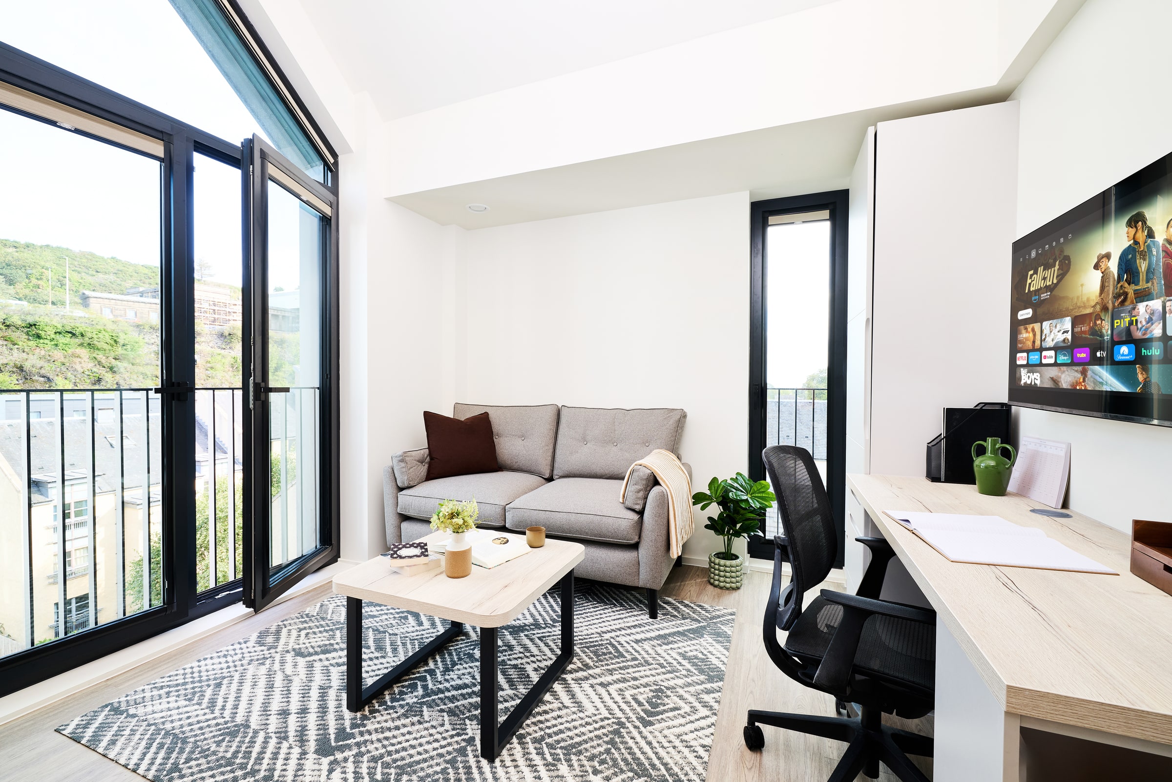 Modern living room with a gray sofa, coffee table, desk, office chair, large windows, indoor plant, and a wall-mounted TV displaying streaming services. Bright natural light fills the space.