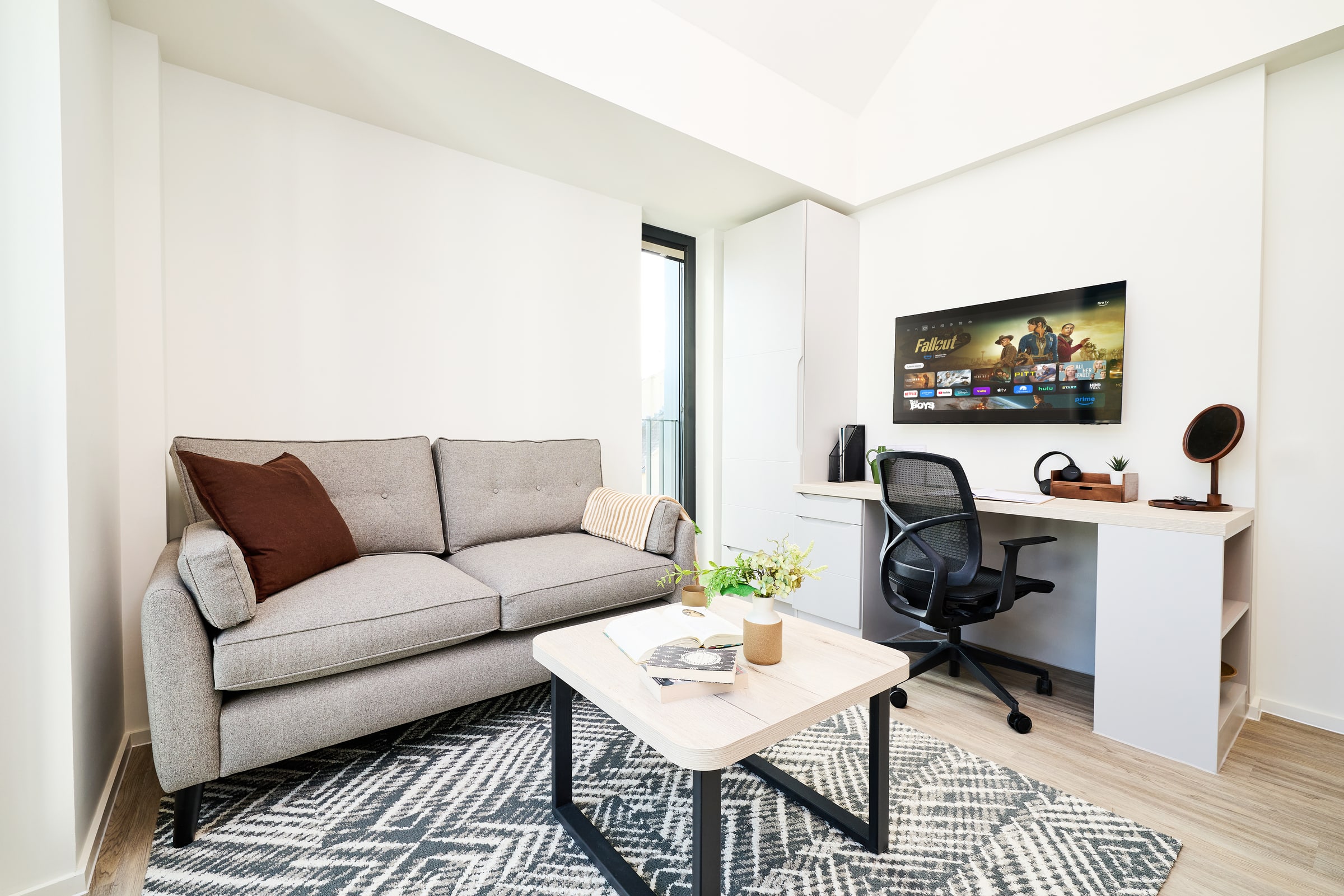 Modern living space with a gray sofa, geometric rug, small coffee table, and a desk setup featuring a computer monitor displaying TV shows. There is also a black office chair and decorative plants.