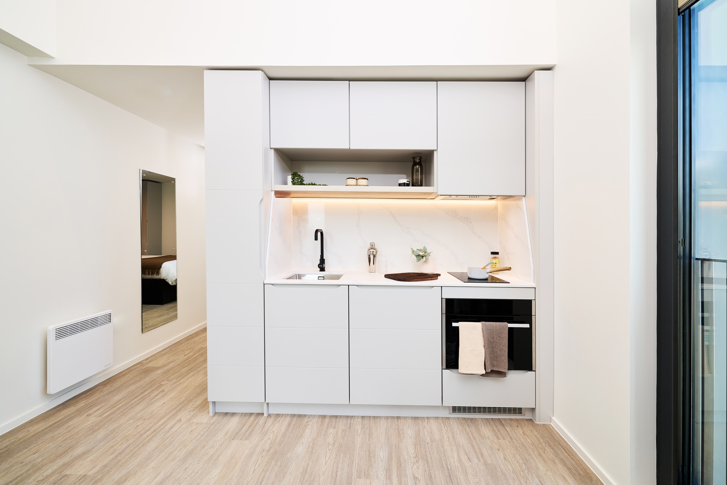 Modern minimalist kitchen with white cabinets, marble backsplash, built-in oven, black faucet, and light wood flooring. A mirror reflects a bed in the adjoining room. Natural light comes from a large window on the right.