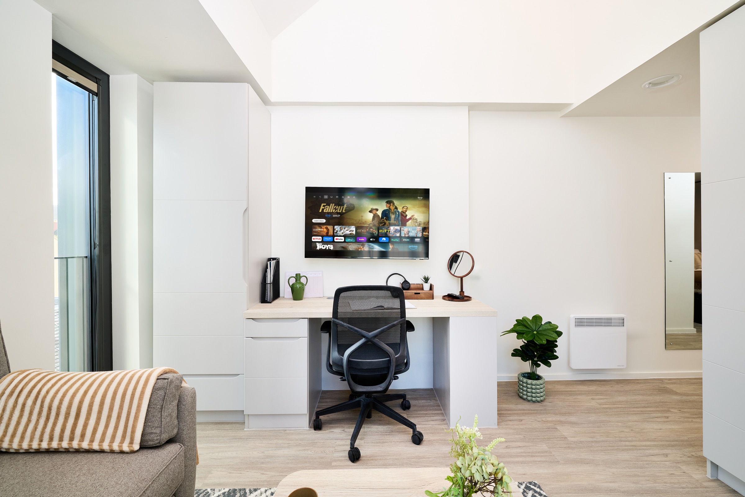 A modern home office with a desk, chair, computer monitor displaying a streaming service, potted plants, a lamp, and a sofa with a striped blanket in a bright, minimalist room.