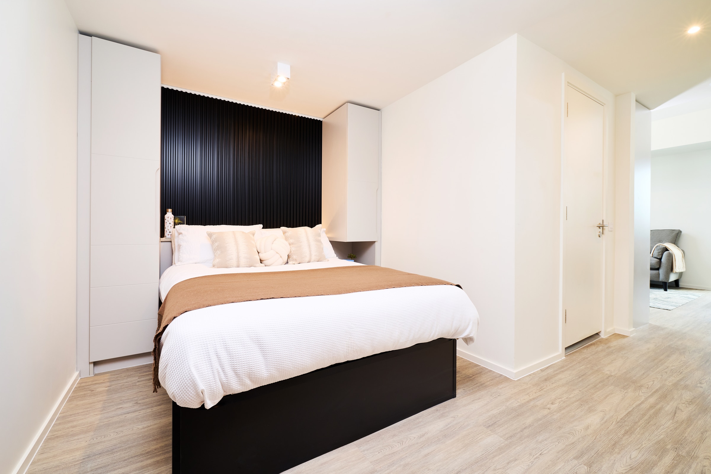 Modern bedroom with a neatly made bed, brown blanket, white pillows, black accent wall, two white wardrobes, light wood floor, and a closed door on the right.