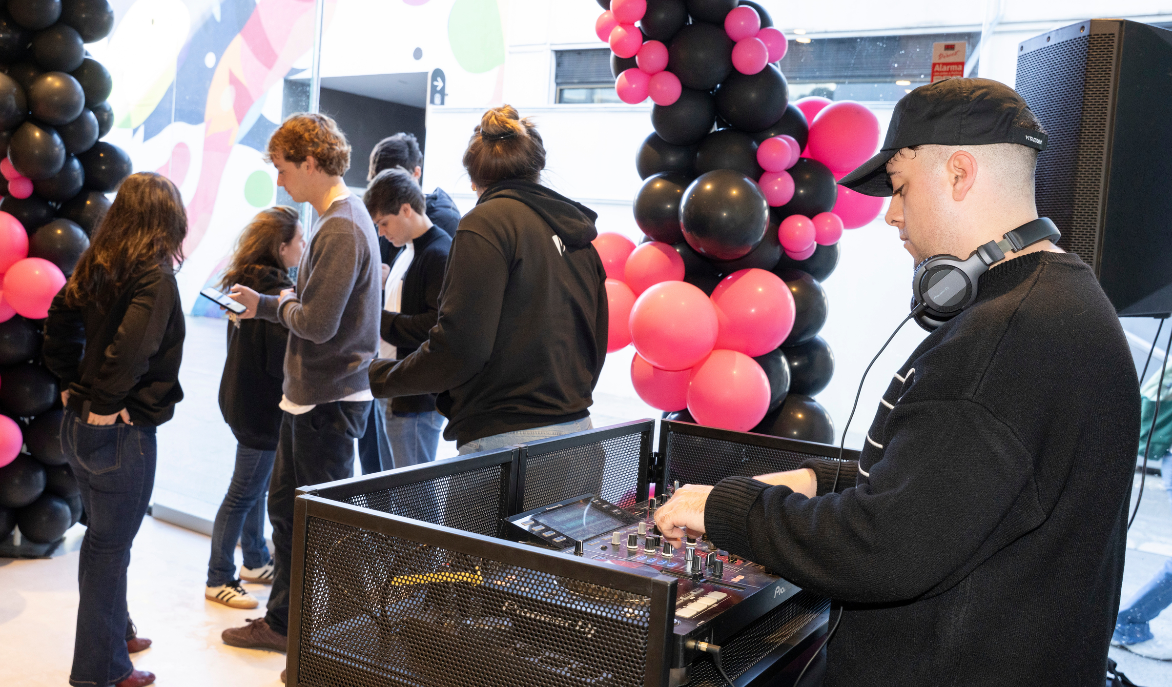 A DJ mixes music at a console near a group of people standing and looking at their phones, with black and pink balloon decorations in the background, setting an energetic vibe for the open day event.