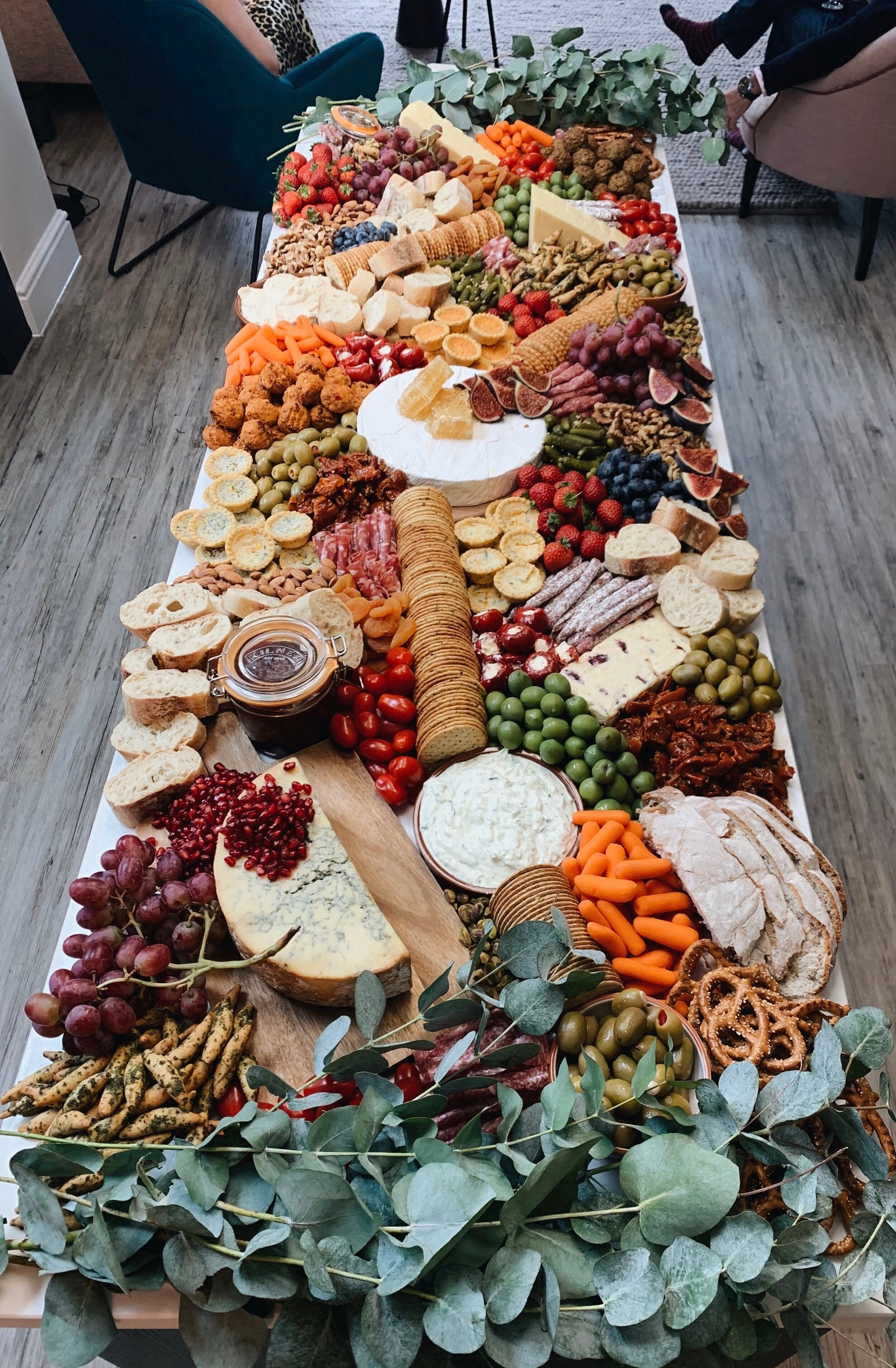 A long grazing table filled with assorted cheeses, crackers, fruits, vegetables, dips, meats, olives, and breads—perfect for your open day—beautifully decorated with greenery on the sides.