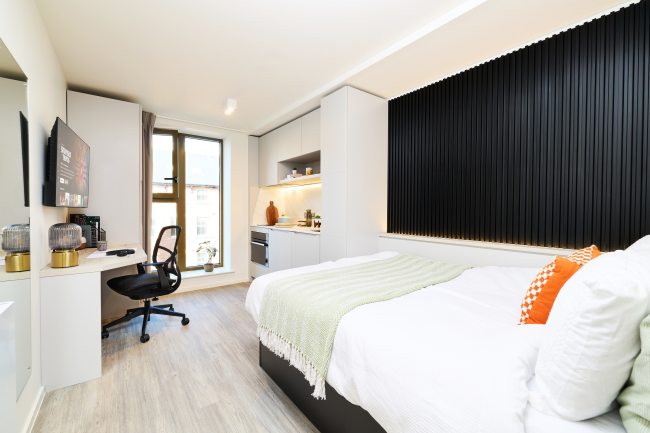 Modern studio apartment with a bed, desk and chair, TV, kitchenette, and large window. The decor is minimalist, featuring white walls, black accents, and light wood flooring.