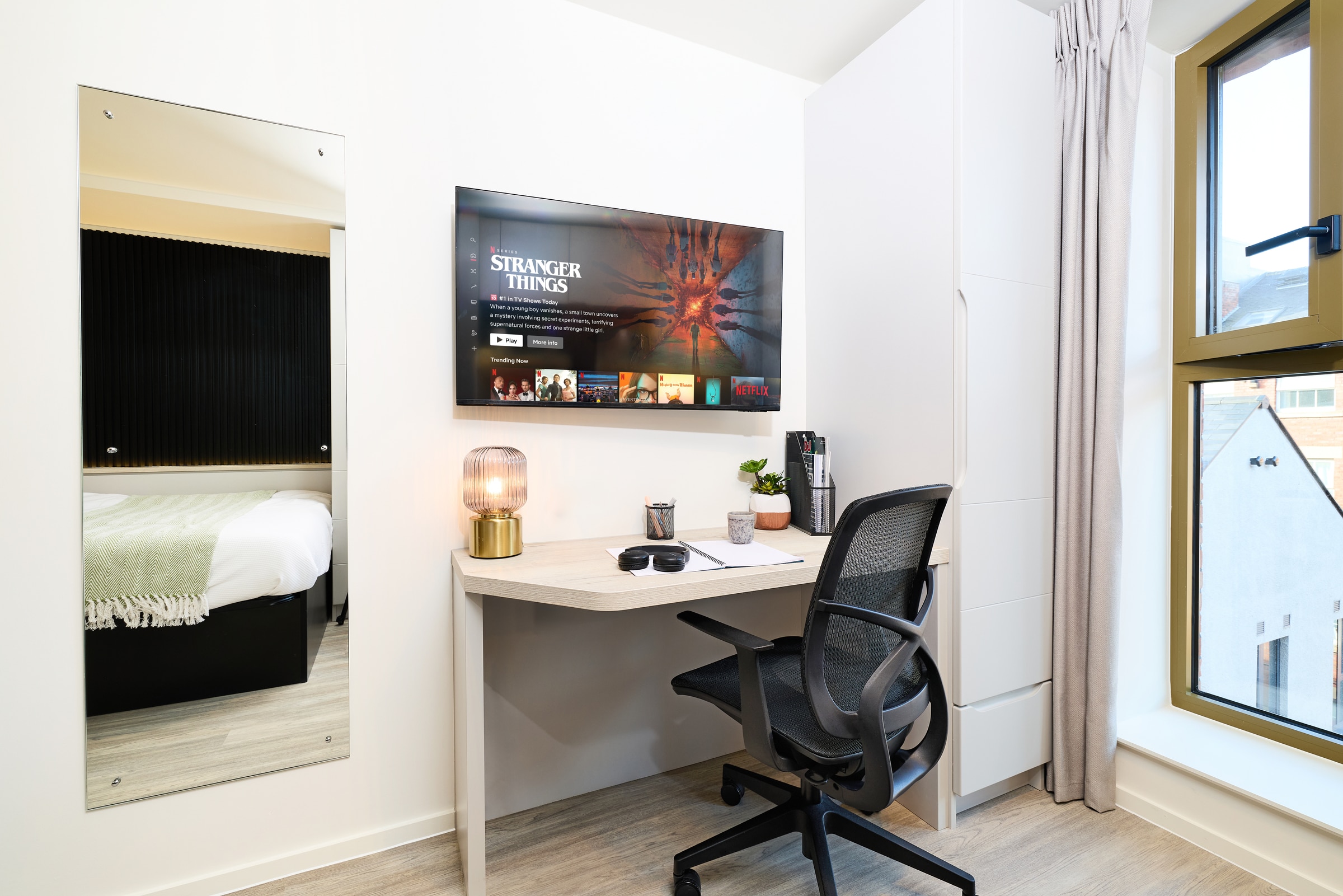 A modern study area with a desk, chair, lamp, and laptop. A TV above the desk displays “Stranger Things.” A bed, large window with curtains, and a tall mirror are also visible.