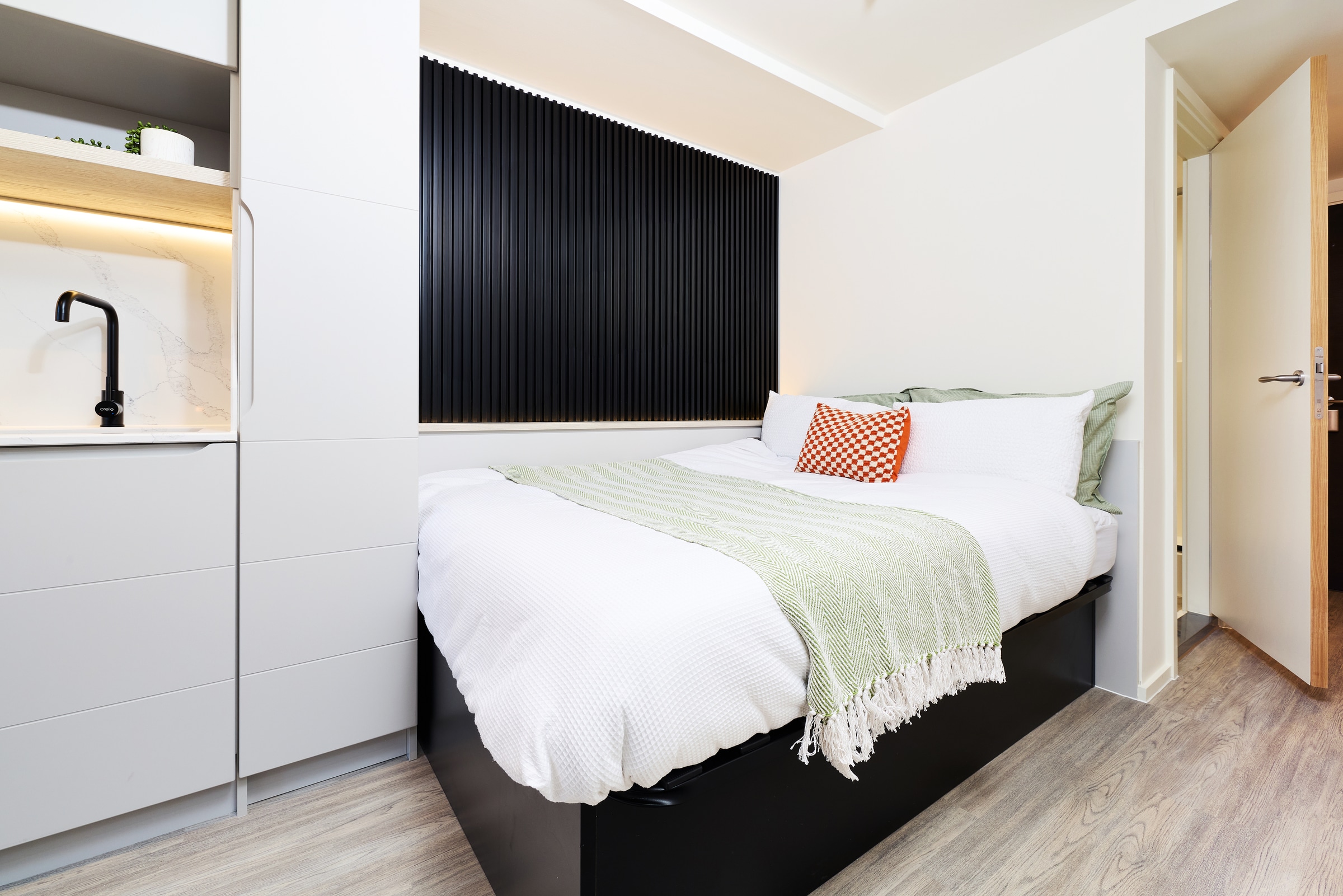 Modern bedroom with a neatly made bed, white linens, green throw, and orange pillow. Black wall panel behind bed, light wood floors, open door, and kitchenette with black faucet on the left. Bright and minimal decor.