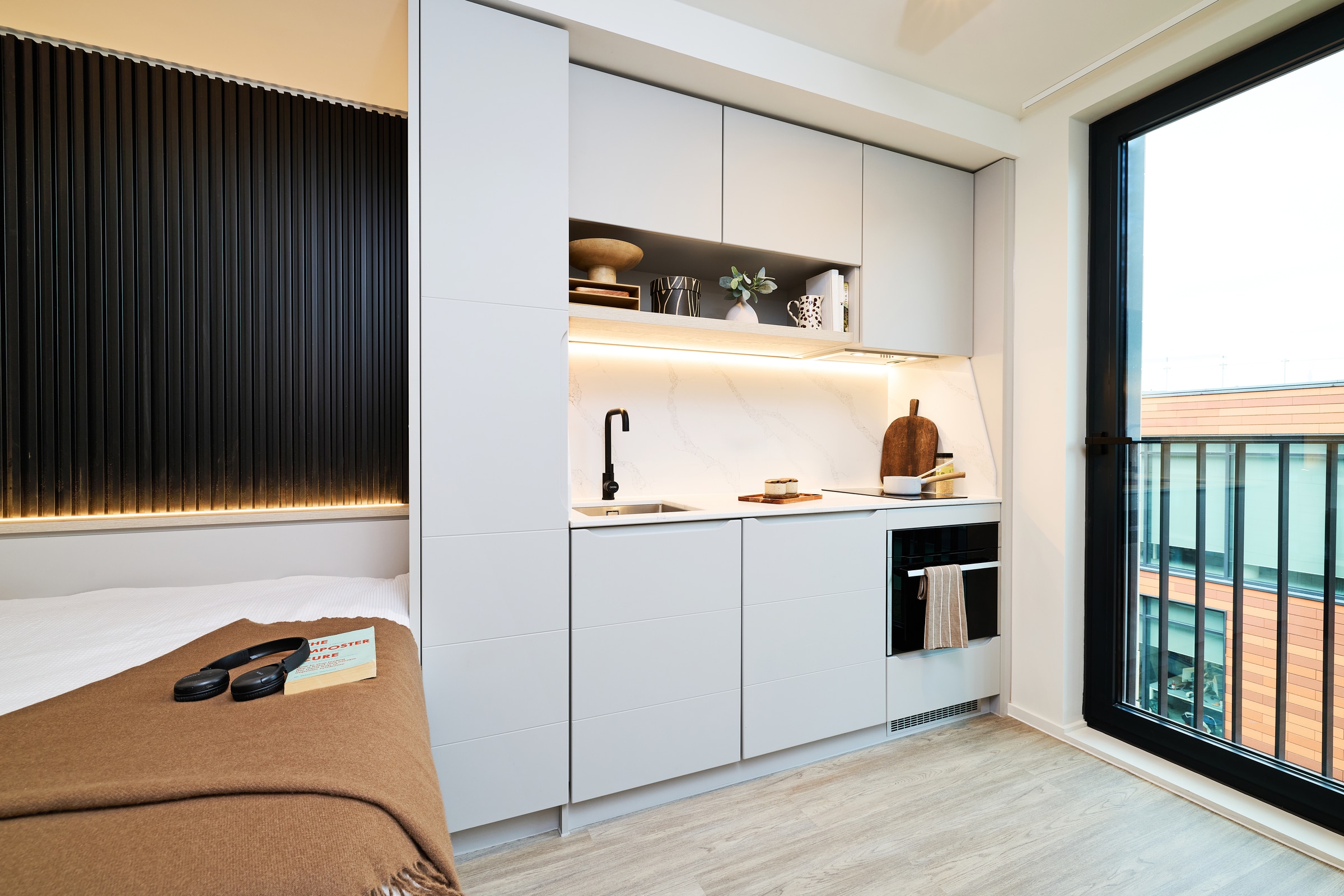 Modern studio apartment with a sleek white kitchenette, black faucet, open shelving, and a bed in the foreground. Large window on the right lets in natural light. Minimalist decor throughout.