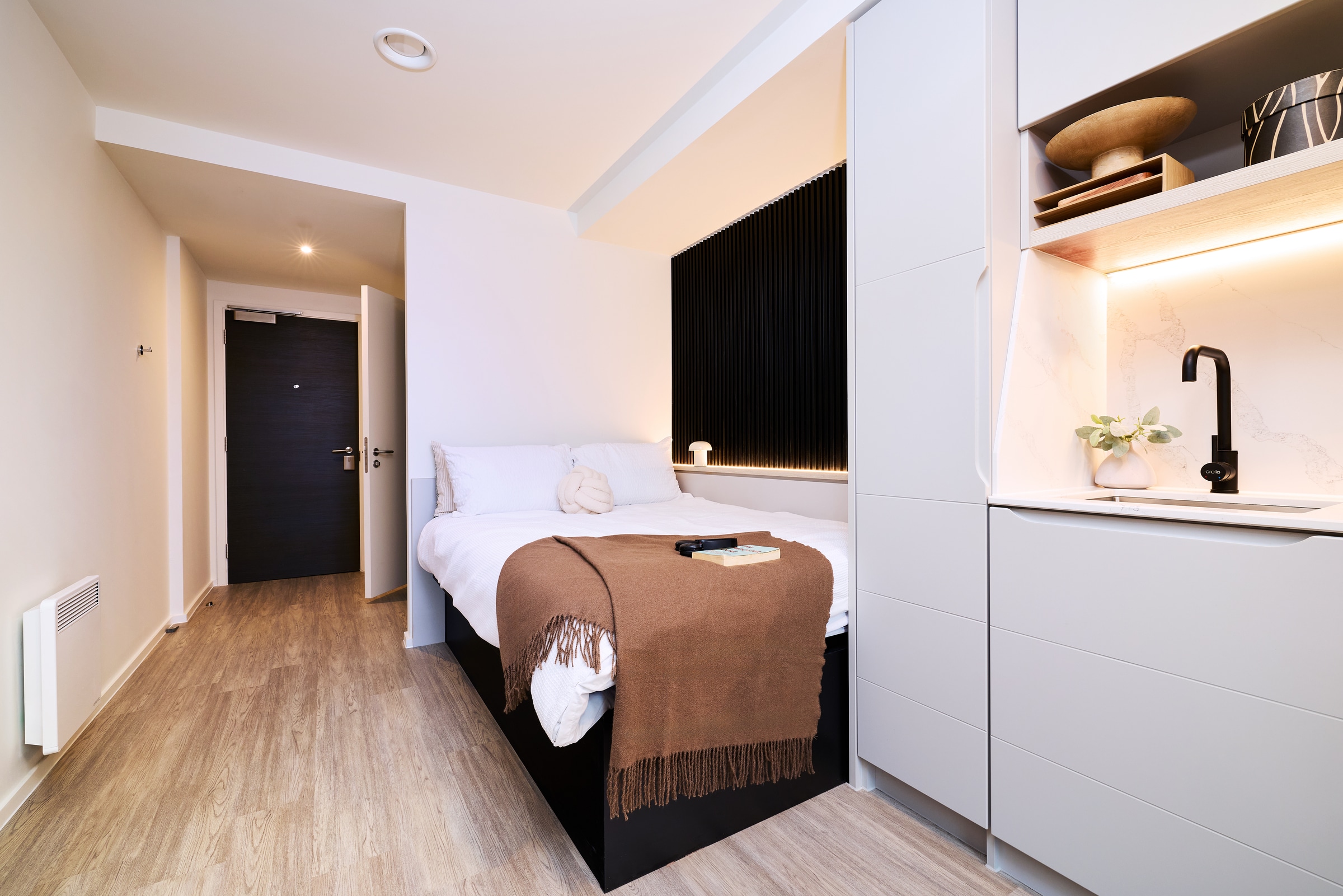 A modern, minimalist studio with a bed, brown blanket, and a book, next to a compact kitchenette with a black sink and light wood floors.