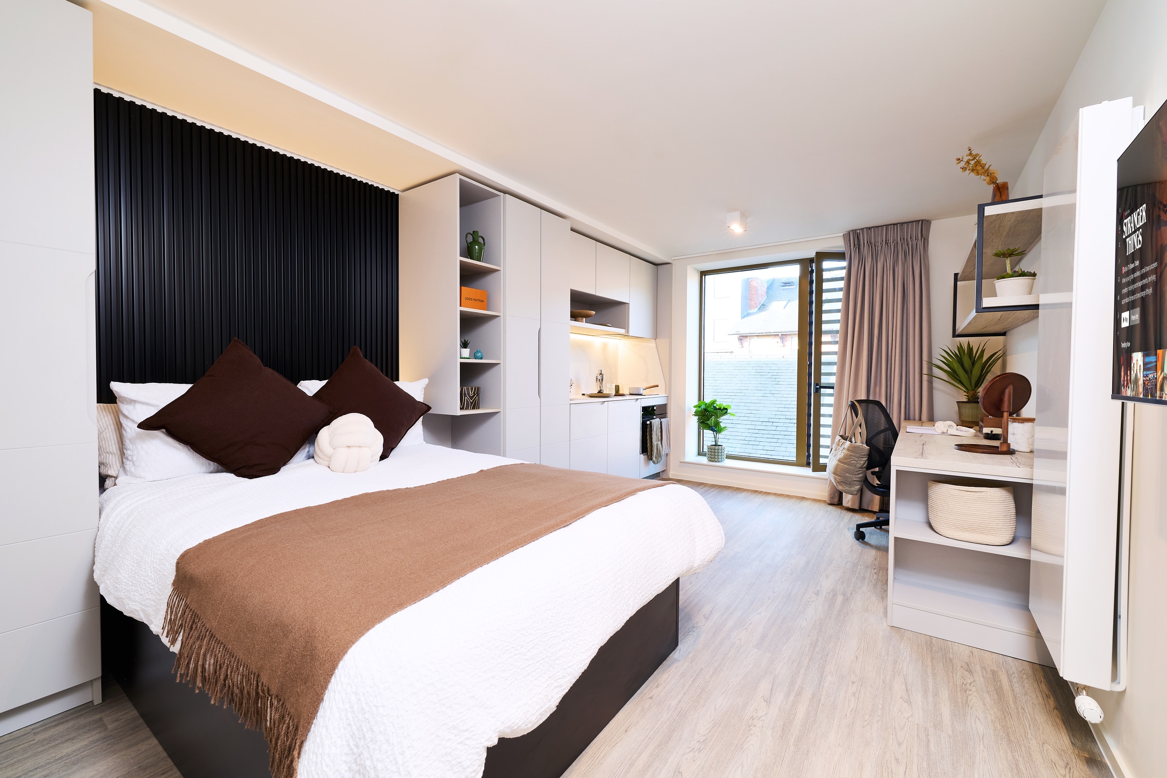 Modern bedroom with a neatly made bed, study desk, chair, open closet, kitchen area, plants, and a large window letting in natural light. Light wood floors and neutral tones create a clean, cozy atmosphere.