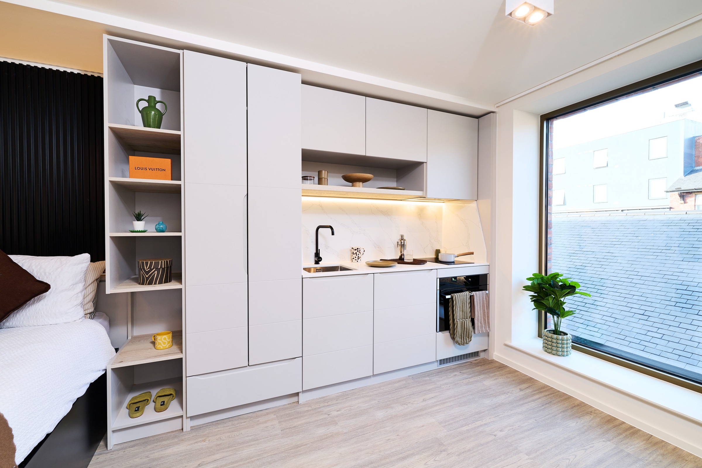 Modern studio kitchen with white cabinets, open shelves with decor, sink, stove, and oven. A large window with a potted plant is on the right, and part of a bed is visible on the left. Light wood floor throughout.