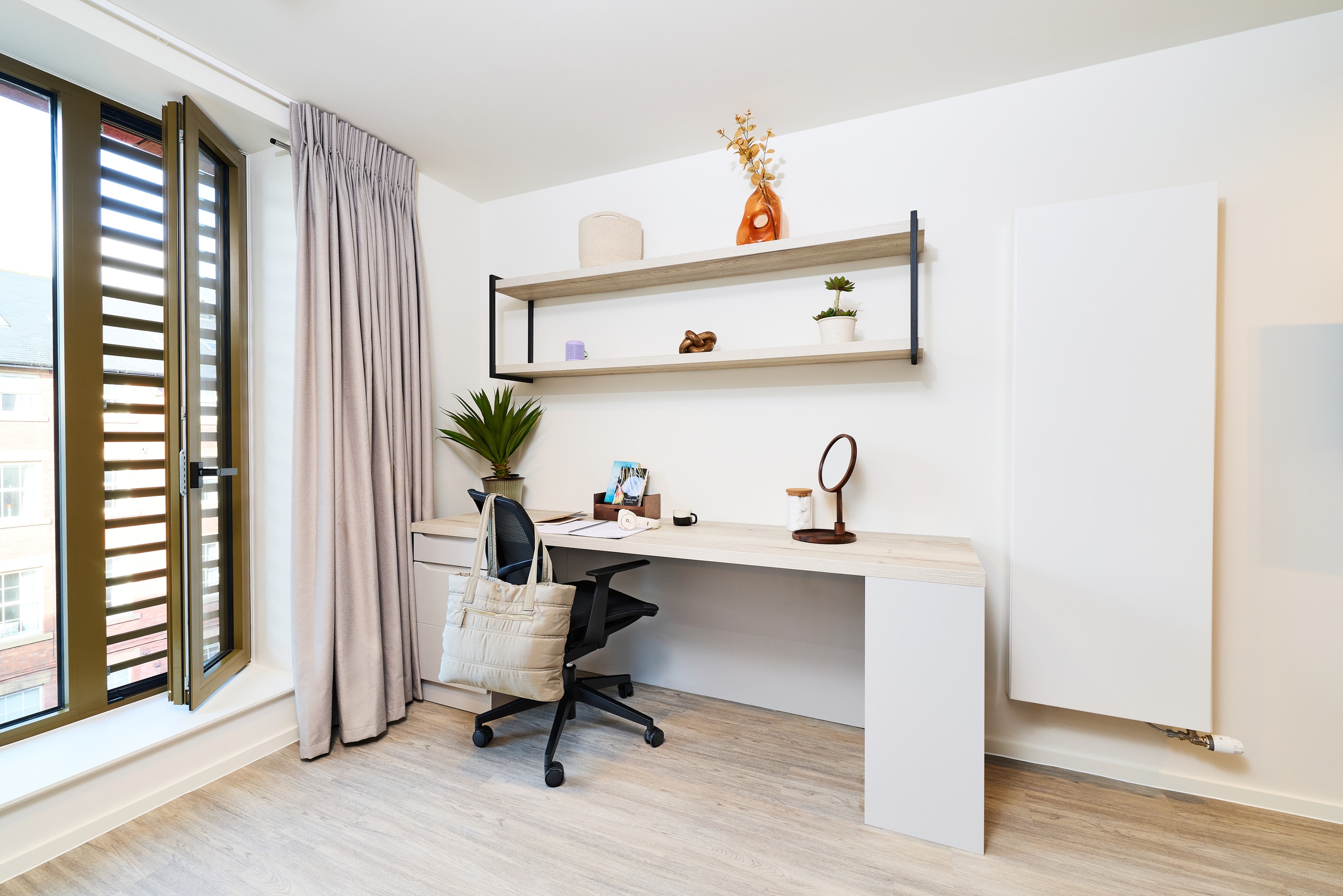 A minimalist home office with a desk, black office chair, open laptop, potted plants, wall shelf with decor, and a window with gray curtains letting in daylight.