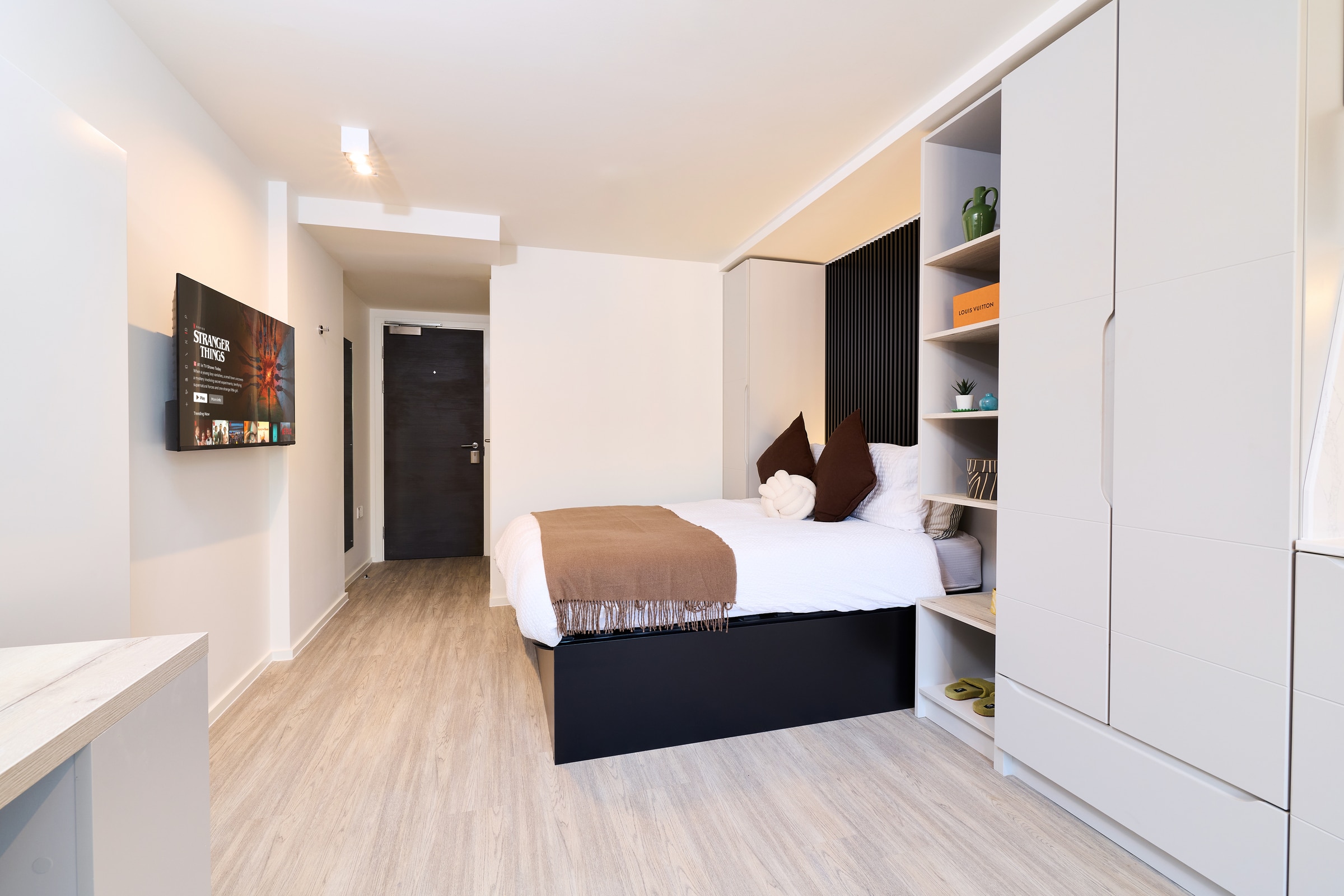 A modern, minimalist bedroom features a neatly made bed with brown and white bedding, built-in shelves and wardrobes, light wood flooring, and a wall-mounted TV opposite the bed.