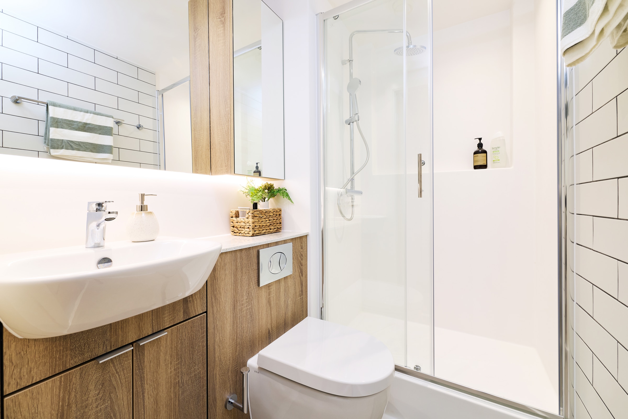 Modern bathroom with wood cabinets, white sink and toilet, glass shower enclosure, white tile walls, and a shelf holding soap, a plant, and a basket. A striped towel hangs on the wall.